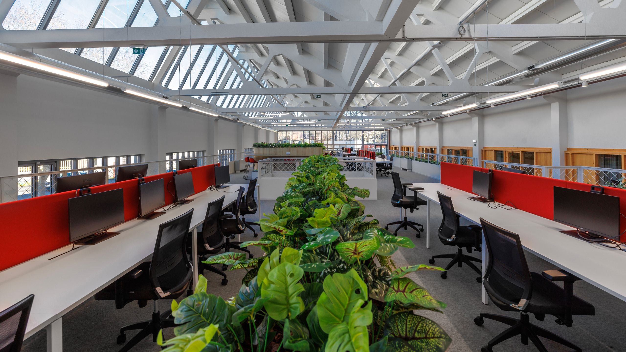 Vista ampla do open space no mezanino com bancadas de trabalho brancas, divisórias vermelhas, plantas centrais e claraboias industriais.