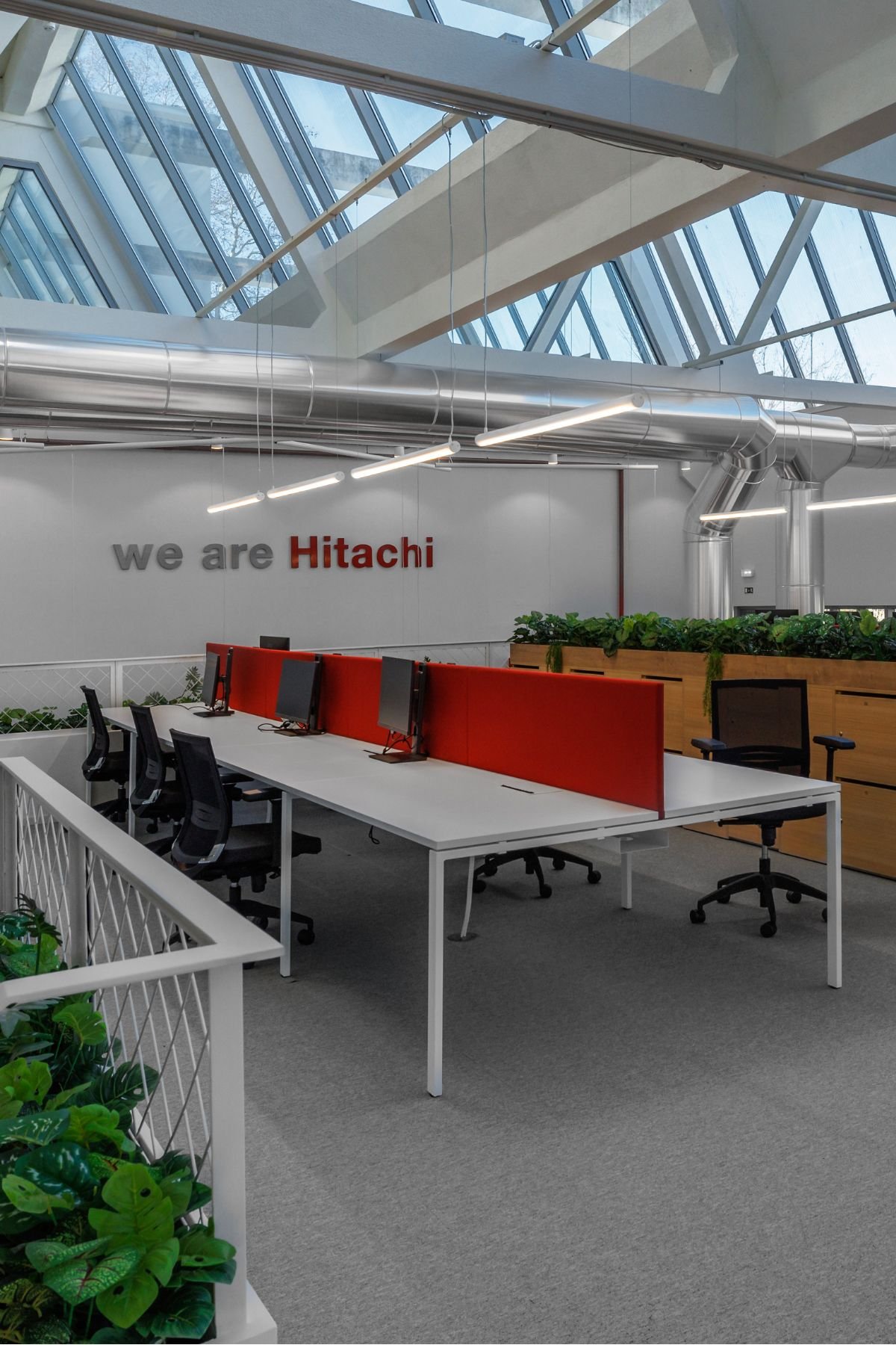 Zona de bancadas de trabalho com painéis divisórios vermelhos e slogan da empresa Hitachi na parede sob teto envidraçado.