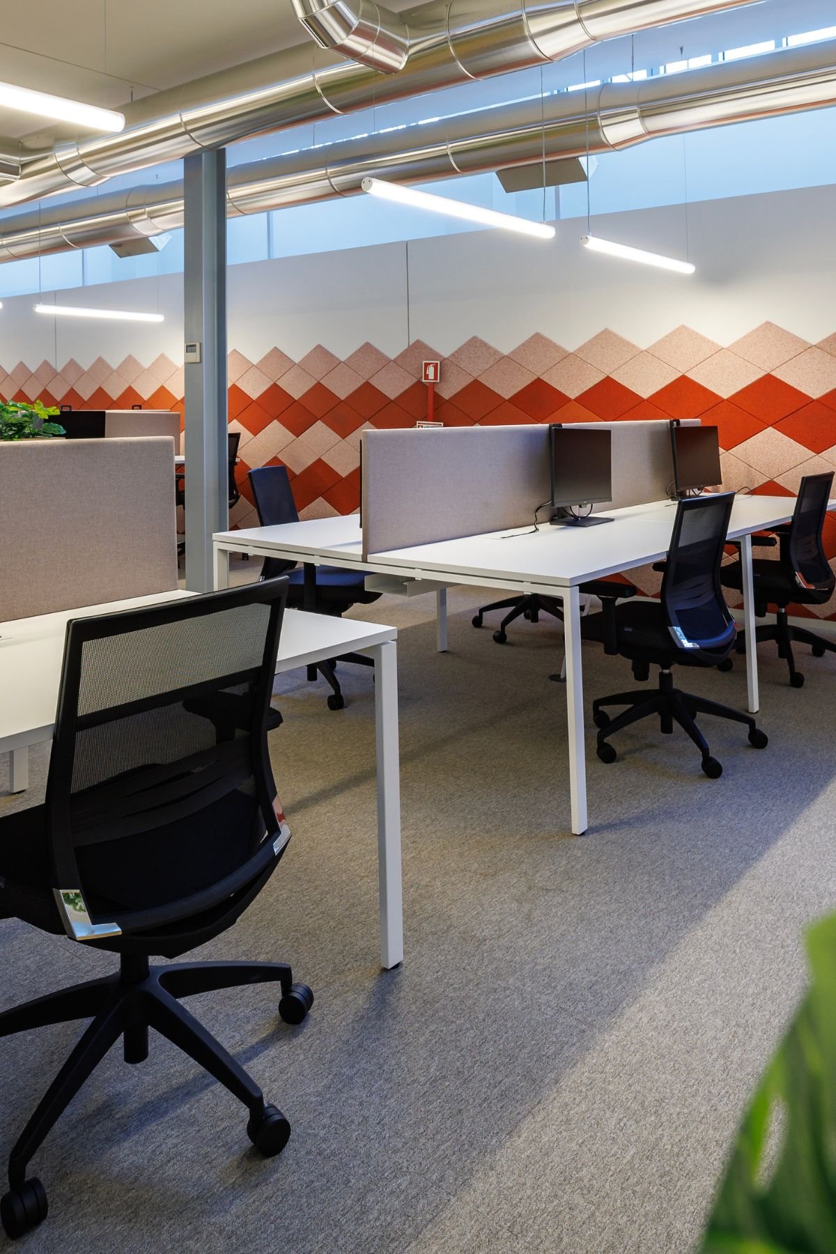 Detalhe de ilhas de trabalho com cadeiras ergonómicas pretas e parede decorativa com revestimento geométrico em tons de vermelho e rosa.