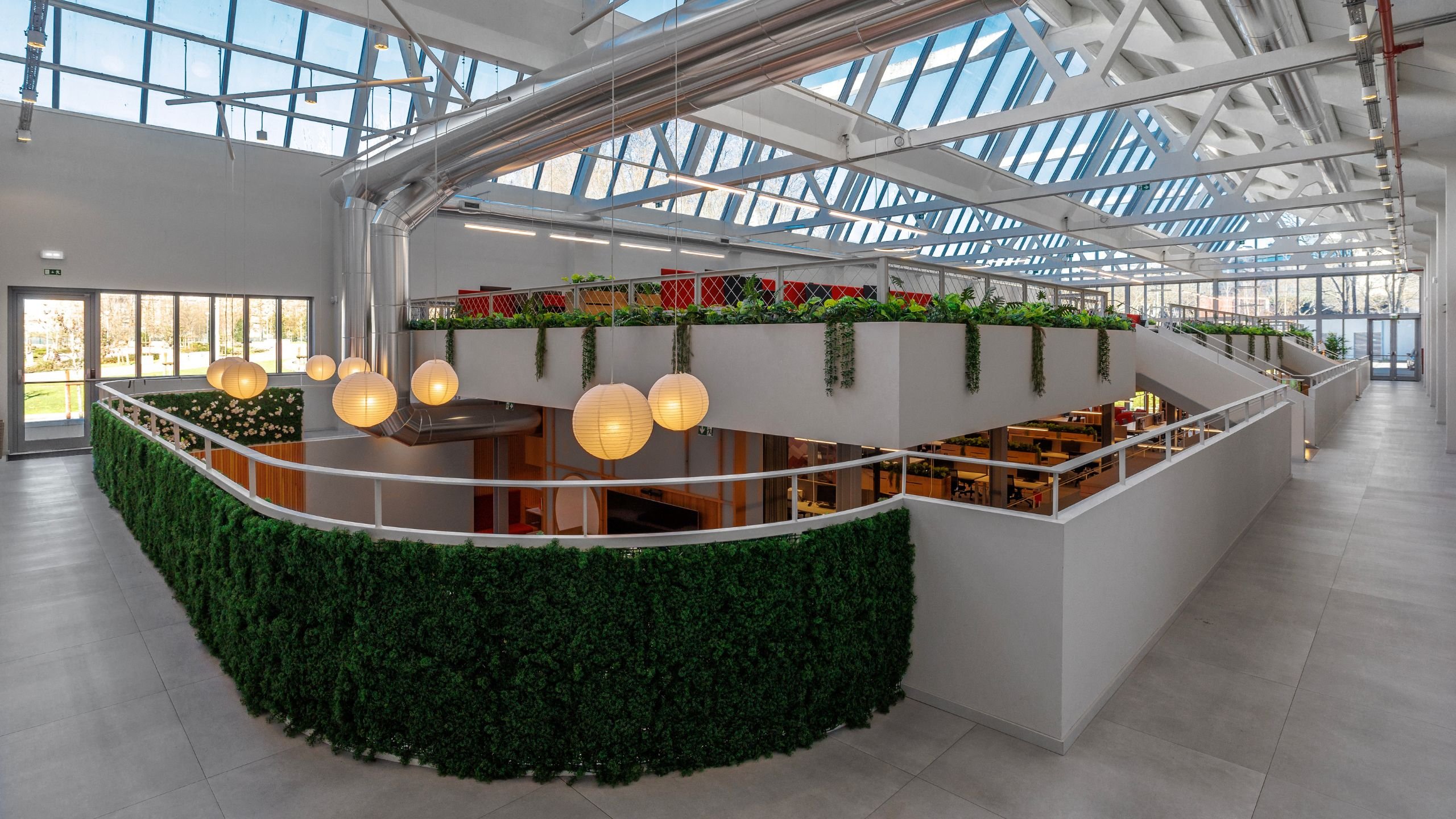 Vista panorâmica do pé-direito duplo do escritório destacando lustres esféricos de inspiração japonesa, plantas suspensas e claraboias.
