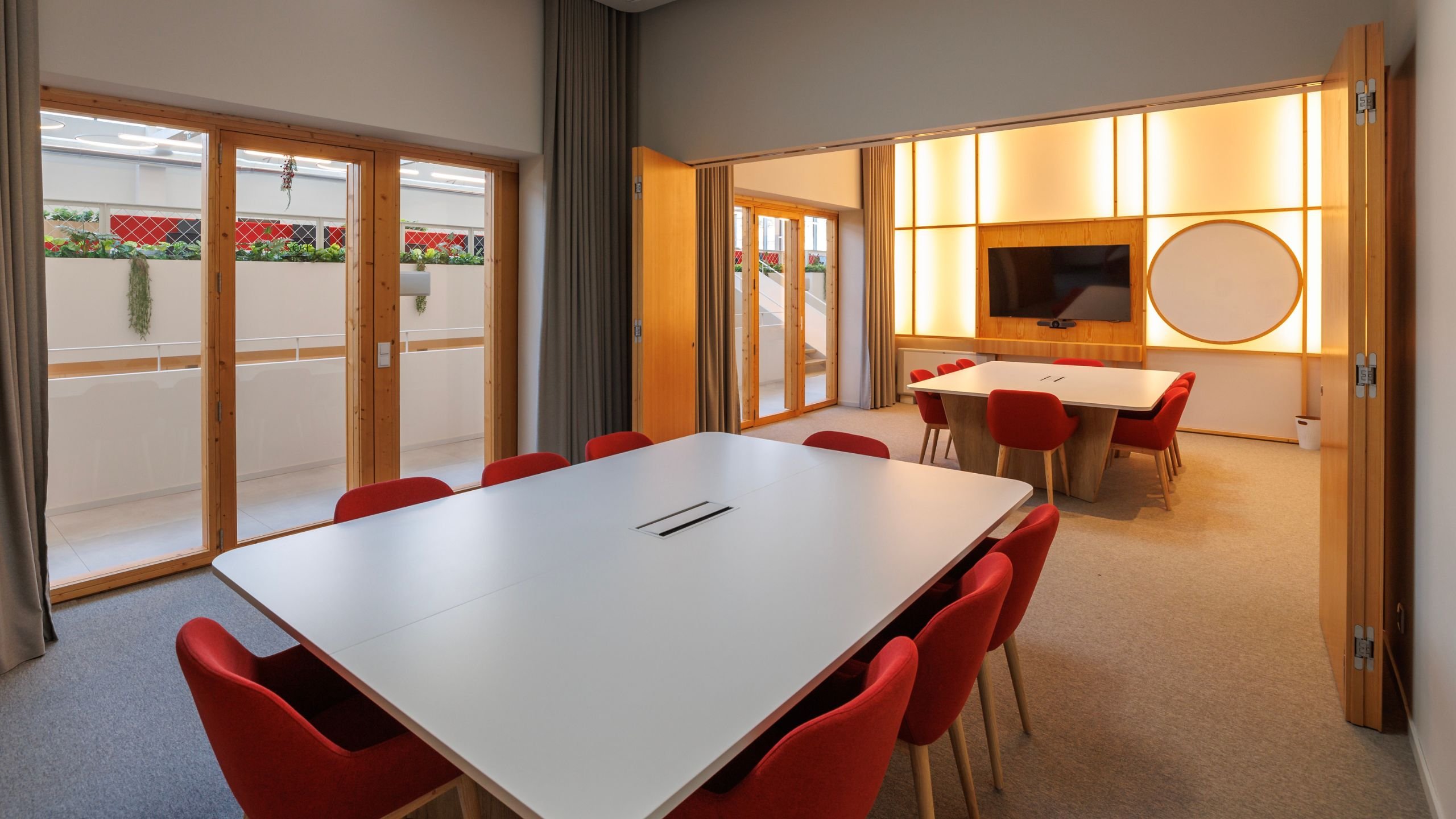 Sala de reuniões ampla com mesas modulares brancas, cadeiras estofadas vermelhas e painel translúcido iluminado ao fundo.