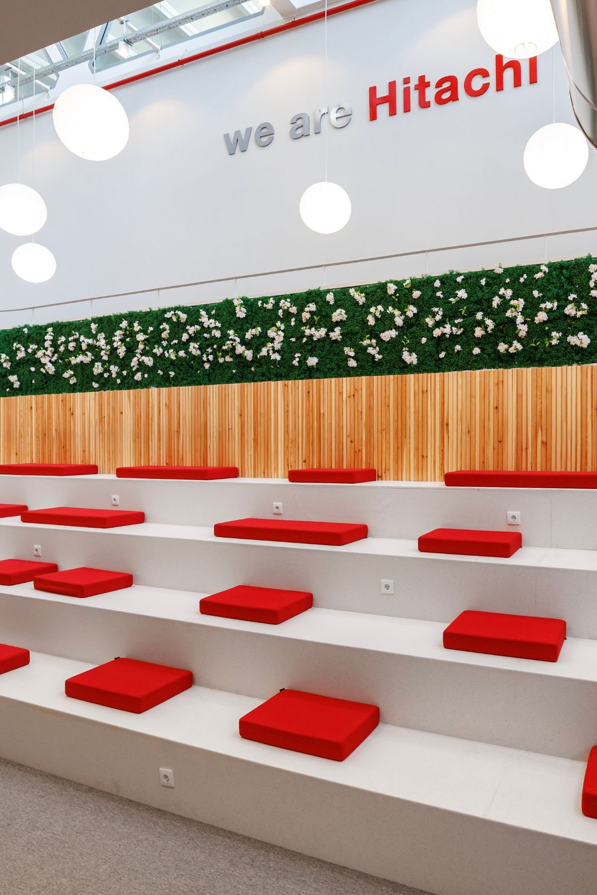 Degraus do anfiteatro em tom claro com almofadas vermelhas dispostas ao estilo tatami, painel em ripas de madeira e sebe floral na parede.