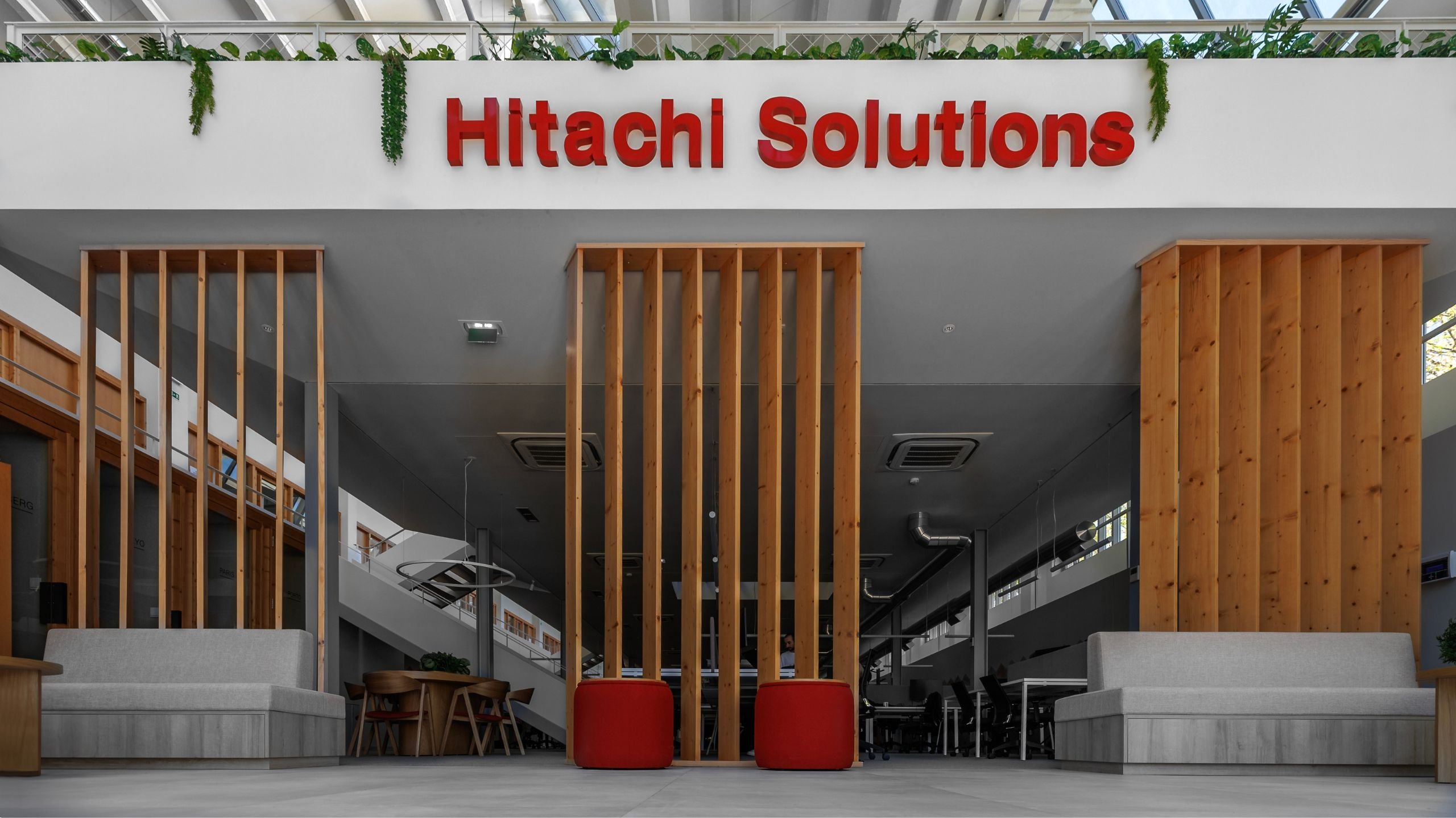 Área de relaxamento e colaboração no hall com sofás cinzentos, pufes vermelhos e divisórias de madeira sob o logótipo da Hitachi Solutions.