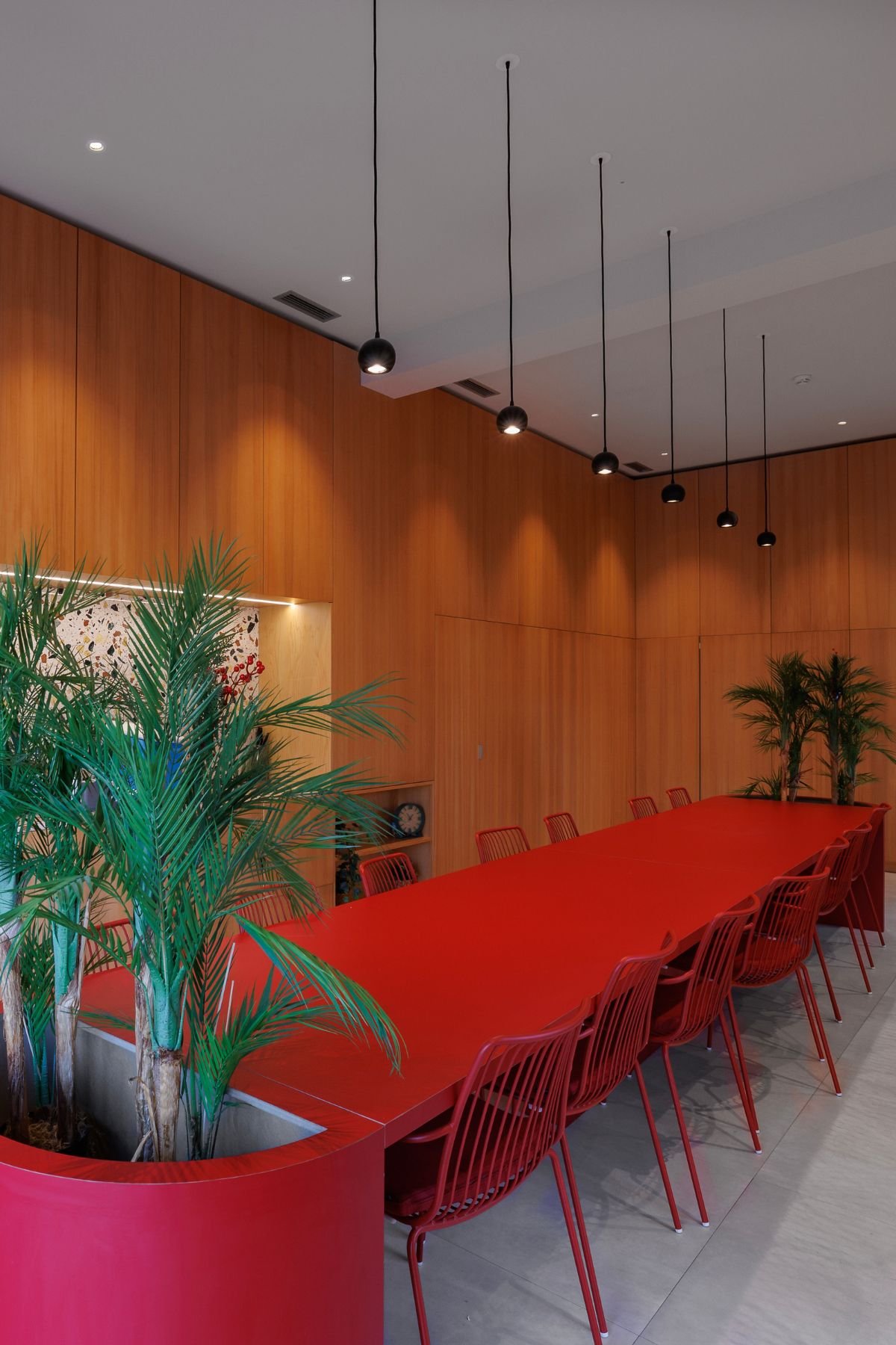 Copa do escritório com mesa central vermelha para refeições partilhadas, plantas decorativas e iluminação moderna suspensa.