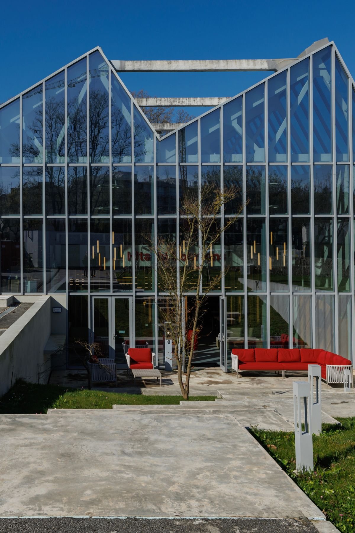 Vista frontal do pátio ao ar livre com elementos naturais, árvore central e entrada em vidro para uma pausa revigorante dos colaboradores.
