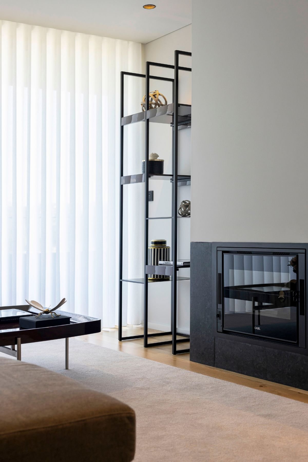 Pormenor de lareira encastrada e estante de metal ao lado de uma mesa de centro e sofa chaise longue, revelando a decoração sala sofisticada numa sala de estar em open space.