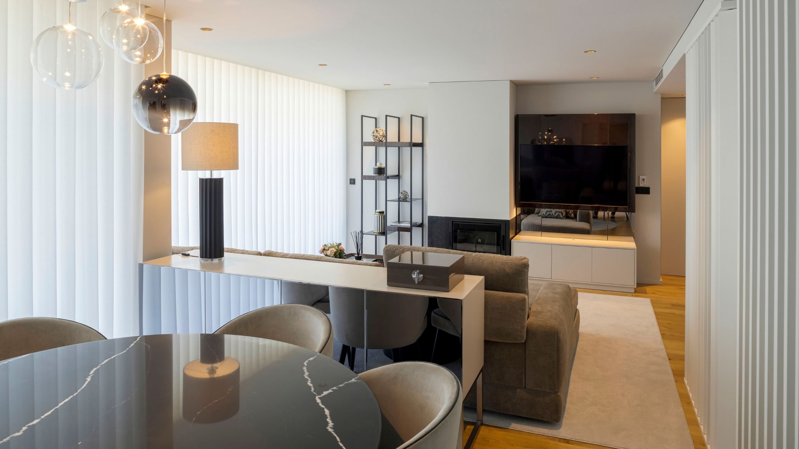Uma vista panorâmica de uma sala de estar e jantar de conceito aberto. No primeiro plano, uma mesa sala de jantar redonda de mármore preto com cadeiras sala de jantar de veludo cinzento claro está posicionada sobre um tapete circular cinzento. Uma luminária pendente de vidro flutua acima da mesa. Ao fundo, uma área de estar com um sofá de linho cinzento, uma consola branca moderna com um candeeiro de mesa e um aparador de média alta, e uma unidade de entretenimento com uma televisão emoldurada por espelhos e um móvel de armazenamento baixo. Uma estante aberta de metal preto exibe objetos de decoração. Este projeto de interior design exemplifica como Aparadores modernos, mesa sala de jantar e cadeira sala de jantar coexistem de forma harmoniosa em uma decoração integrada.