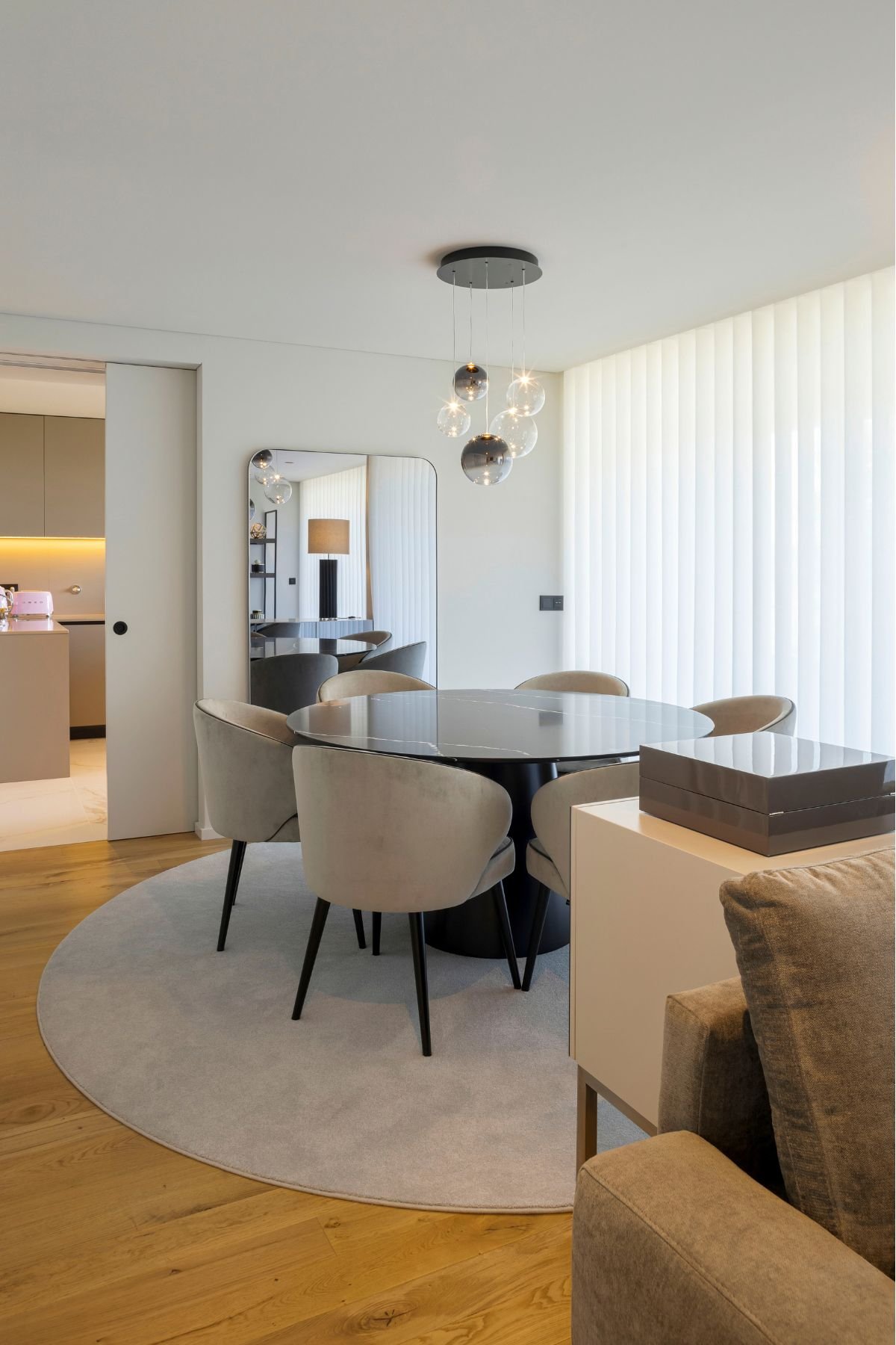 Uma vista panorâmica de uma sala de jantar de conceito aberto. Ao centro, uma mesa sala de jantar redonda de mármore preto com cadeiras sala de jantar de veludo cinzento claro está posicionada sobre um tapete circular cinzento. Ao fundo, um espelho de corpo inteiro emoldurado por metal preto reflete uma prateleira aberta. À direita, um aparador branco moderno com tampo de vidro e caixas decorativas oferece armazenamento. Uma luminária pendente de vidro flutua acima da mesa. Este espaço de interior design moderno combina Aparadores modernos com uma decoração minimalista e sofisticada.