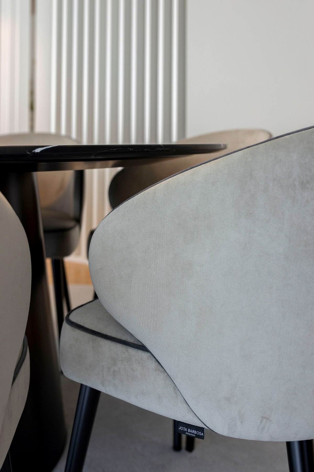 Um plano de detalhe focado nas costas e apoio de braço curvo de uma cadeira sala de jantar de veludo cinzento claro com debrum preto e pés pretos de madeira, que inclui uma pequena etiqueta de metal 'JOTA BARBOSA INTERIORS'. Ao fundo, vê-se uma divisória de ripado branco vertical e outra cadeira semelhante. Esta cadeira sala de jantar é parte de um projeto de interior design focado em mobiliário personalizado e sofisticado, combinando perfeitamente com Aparadores modernos e uma mesa sala de jantar redonda de mármore preto visível à esquerda.