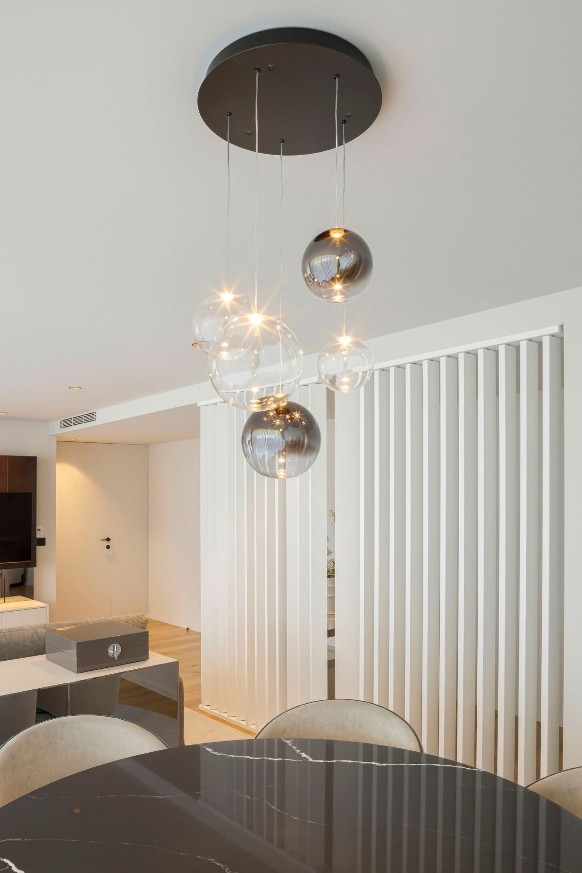 Um conjunto de luminárias pendentes de vidro esférico transparente e prateado espelhado flutua sobre uma mesa sala de jantar redonda de mármore escuro com veios brancos, complementada por cadeiras sala de jantar de veludo cinzento claro com debrum preto. Ao fundo, uma divisória de ripado branco vertical e um corredor levam a um espaço de interior design moderno. A decoração de luxo inclui um aparador espelhado com uma caixa decorativa. Esta imagem demonstra como Aparadores modernos e iluminação de destaque se unem em uma decoração coesa.