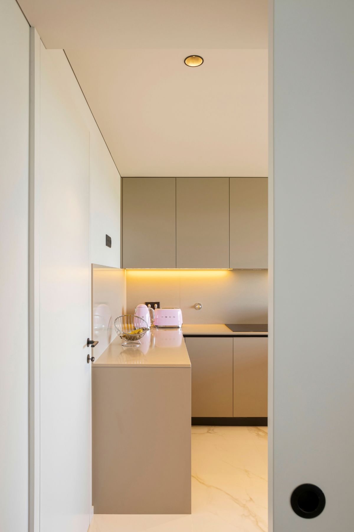 Vista através de um batente de porta de uma cozinha moderna minimalista com armários bicolores em branco e bege acinzentado, bancadas de quartzo branco e eletrodomésticos SMEG cor-de-rosa (chaleira e torradeira). A iluminação LED sob os armários superiores cria um ambiente acolhedor. Este design de cozinha reflete os princípios de interior design aplicados a Aparadores modernos para armazenamento funcional. A decoração clean é complementada por um piso de mármore branco com veios cinzentos. Embora a imagem foque na cozinha, o estilo pode ser estendido para a escolha de uma mesa sala de jantar e cadeira sala de jantar coordenadas.