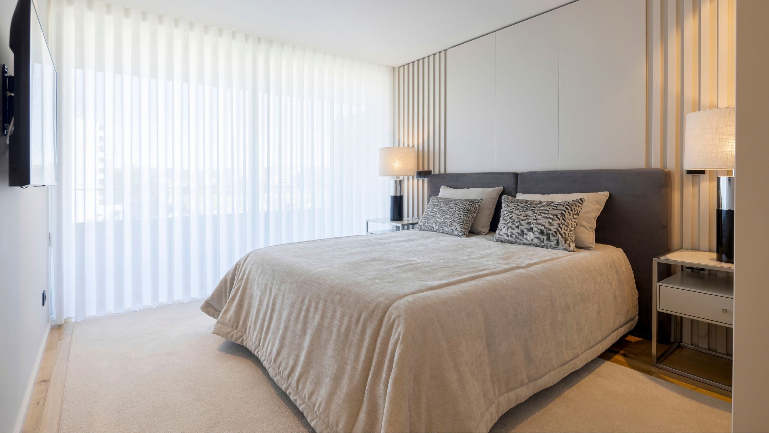 Uma visão panorâmica de um quarto de casal decorado moderno, iluminado por luz natural que atravessa uma cortina branca de privacidade. A cama está vestida com colchas beges e almofadas com padrões geométricos, apoiada em uma cabeceira cinzenta estofada. As paredes laterais apresentam uma textura de ripas de madeira clara, exemplificando o interior design minimalista desta casa moderna. Mesas de cabeceira espelhadas com candeeiros de mesa pretos e abajures beige flanqueiam a cama. Uma televisão de ecrã plano está montada na parede oposta. Esta decoração de quarto foca no conforto e estética.