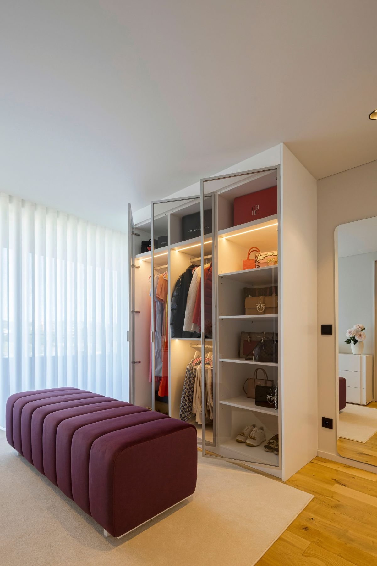 Armário iluminado em closet aberto com portas de vidro num amplo walking closet com toucador com espelho ao fundo, exemplo sofisticado de um dream closet.