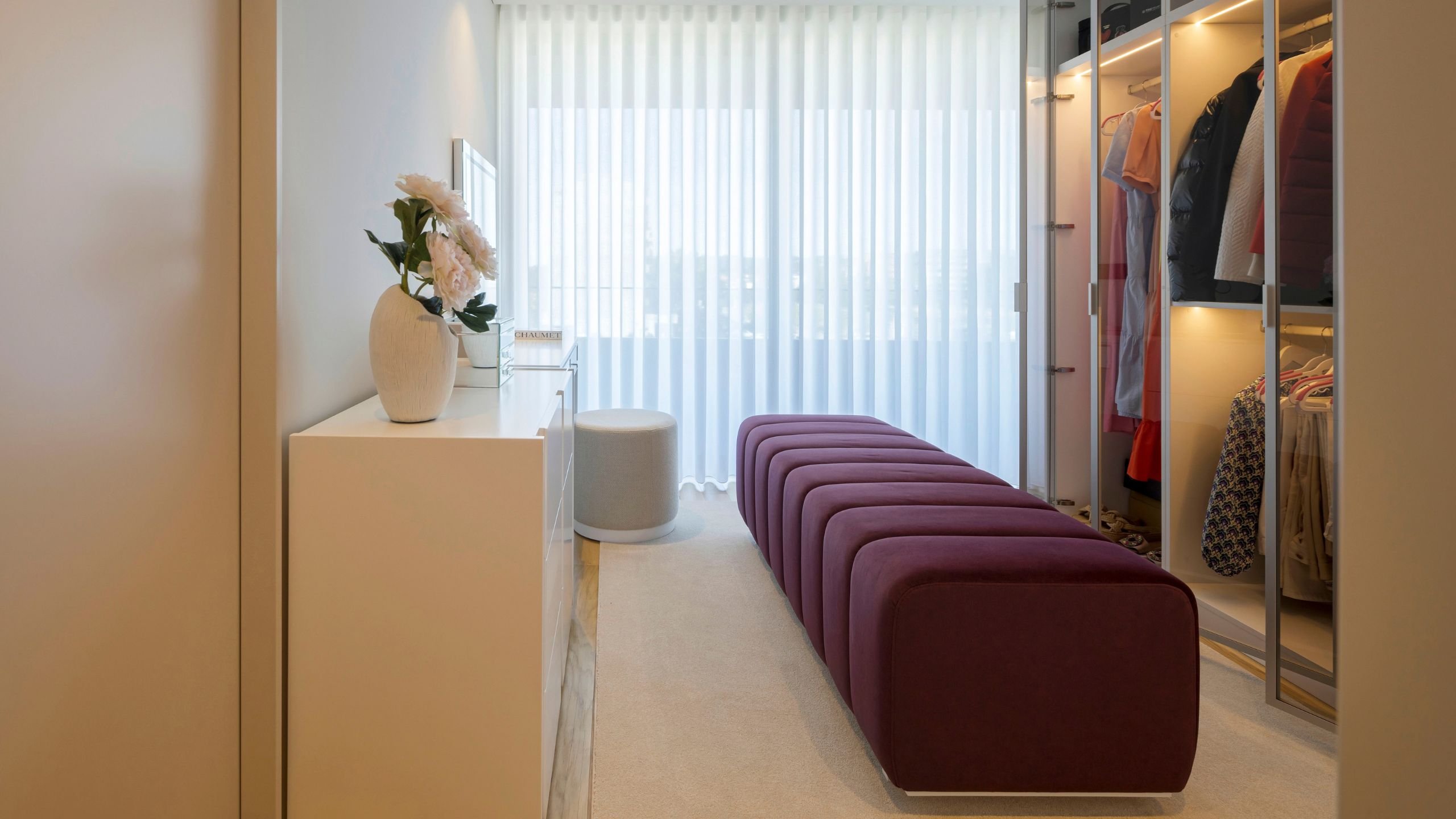Vista de um walking closet luminoso com banco estofado, armários de closet aberto e zona de toucador com espelho, concretizando um verdadeiro dream closet moderno.