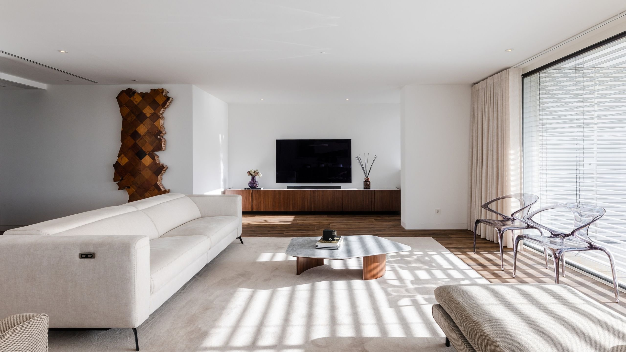 Perspetiva da sala de estar banhada por luz natural, destacando o sofá elétrico branco e as cadeiras transparentes junto à janela.