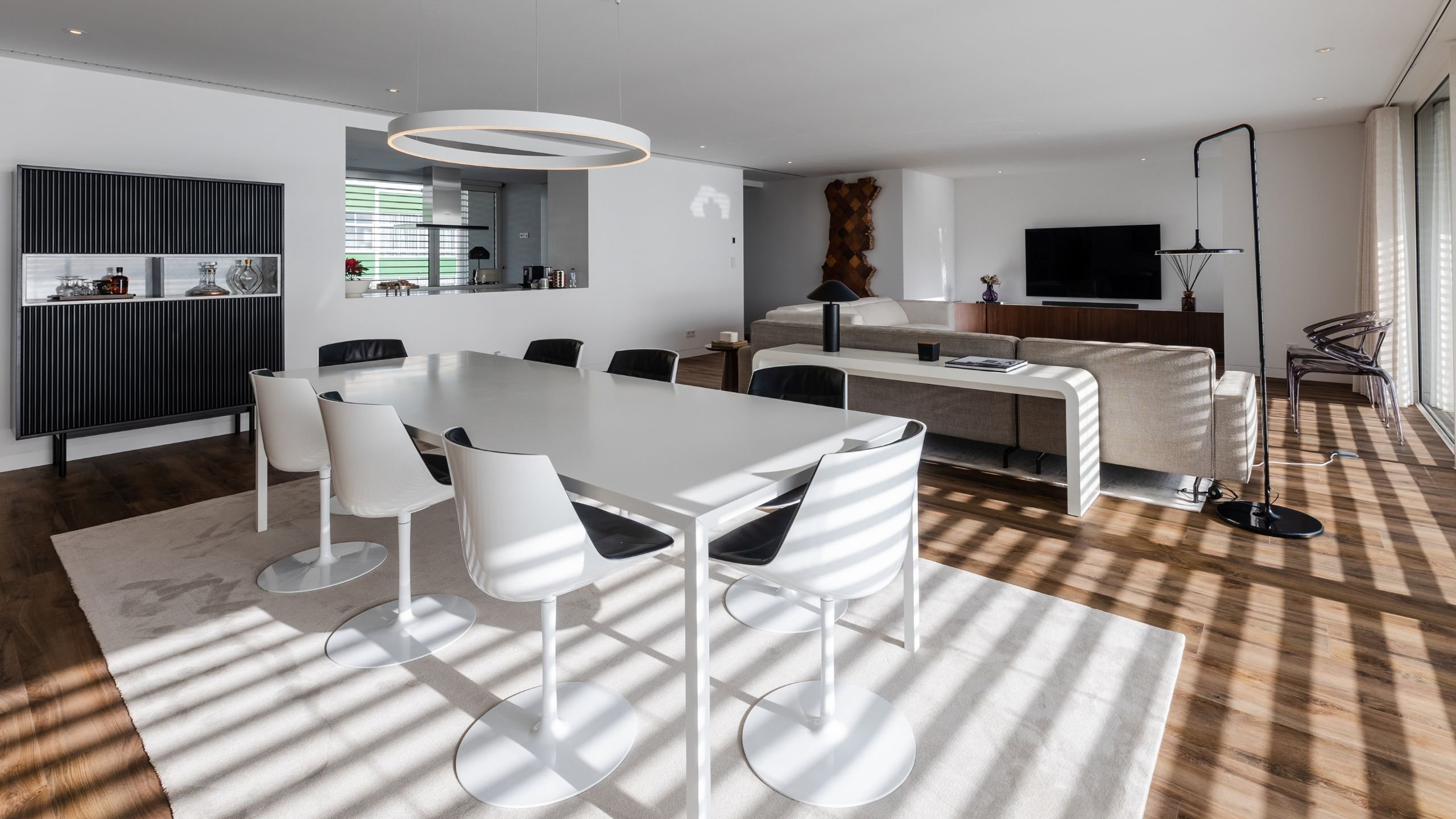 Vista geral da sala de jantar integrada, em plano aberto, realçando a mesa branca sobre o tapete e o candeeiro suspenso circular.