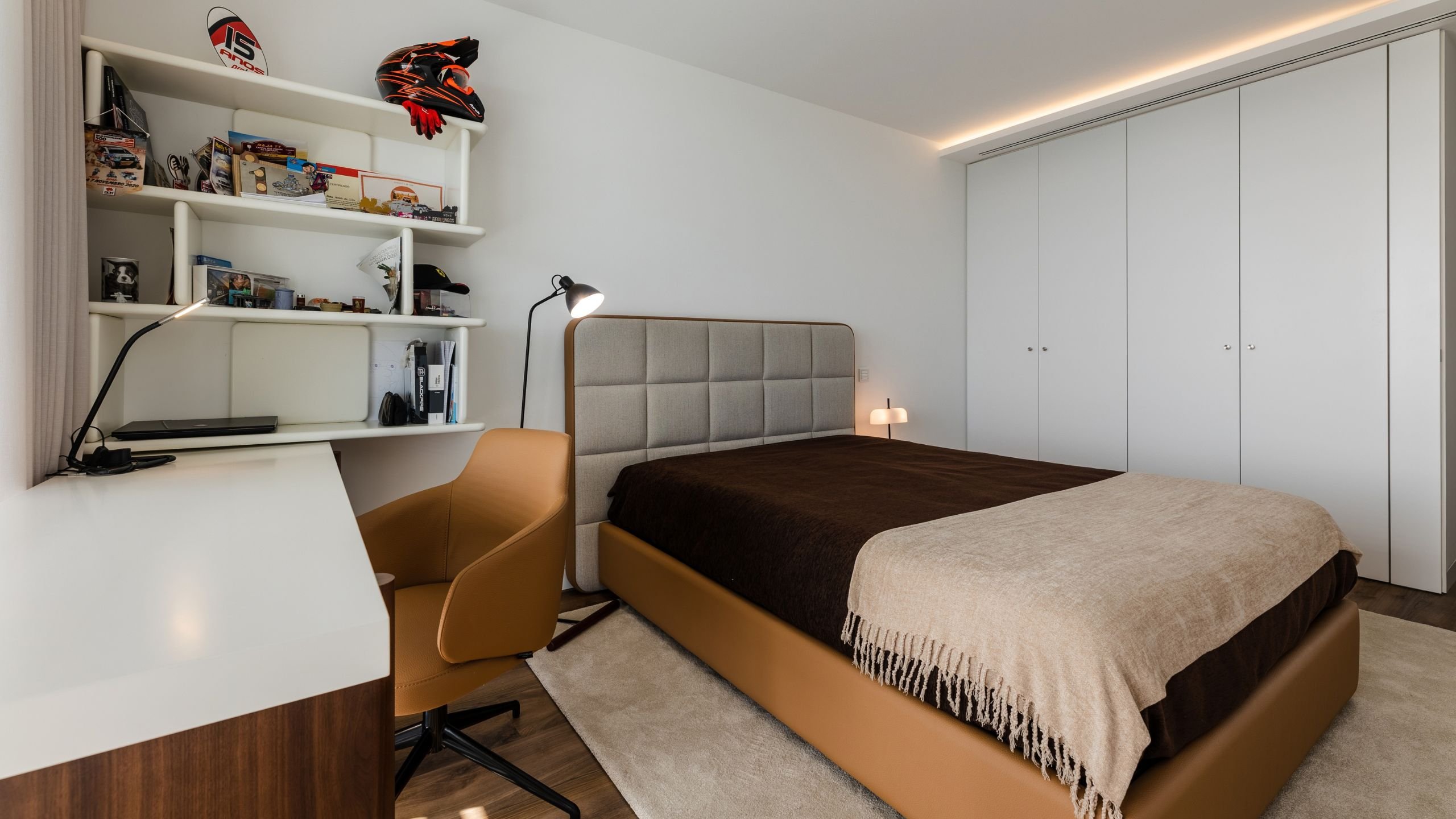 Vista geral do quarto moderno com cama de casal revestida em pele, zona de estudo com secretária e estante ao fundo.
