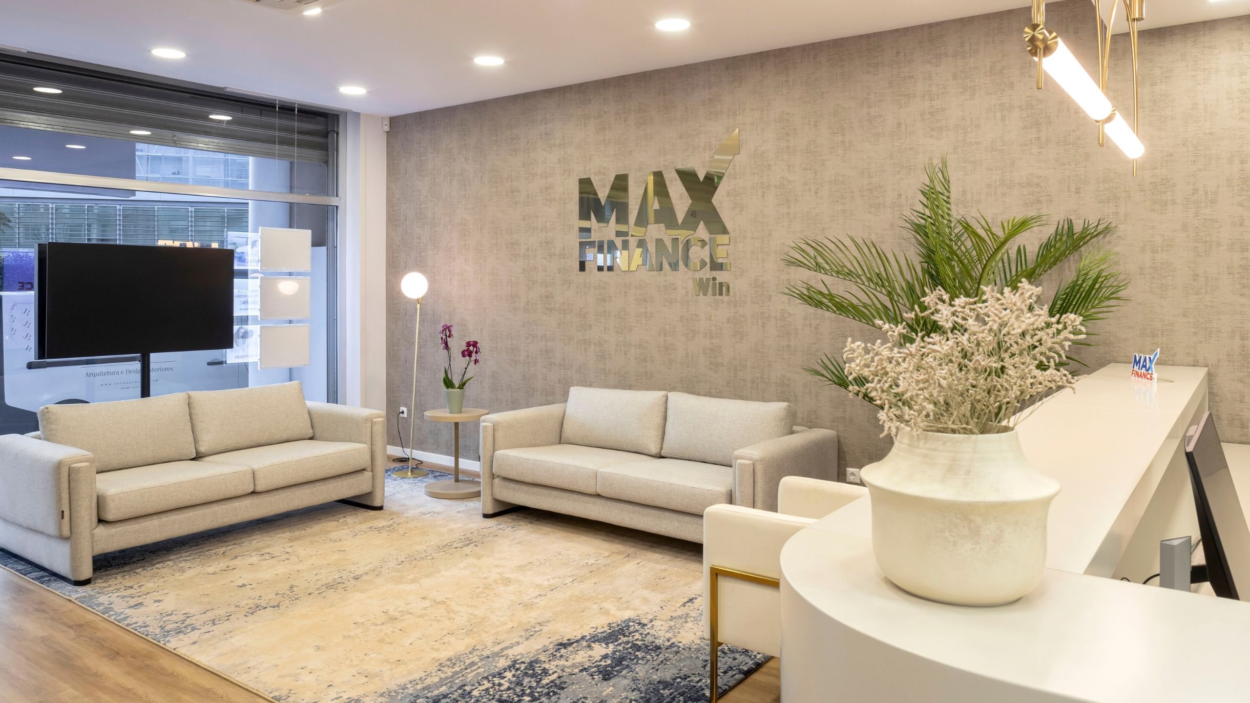 Visão geral da zona de entrada da Imobiliária Max Finance Win, com receção, sofás em tecido cru, papel de parede texturizado bege com o logotipo dourado e tapete com detalhes em azul-marinho.