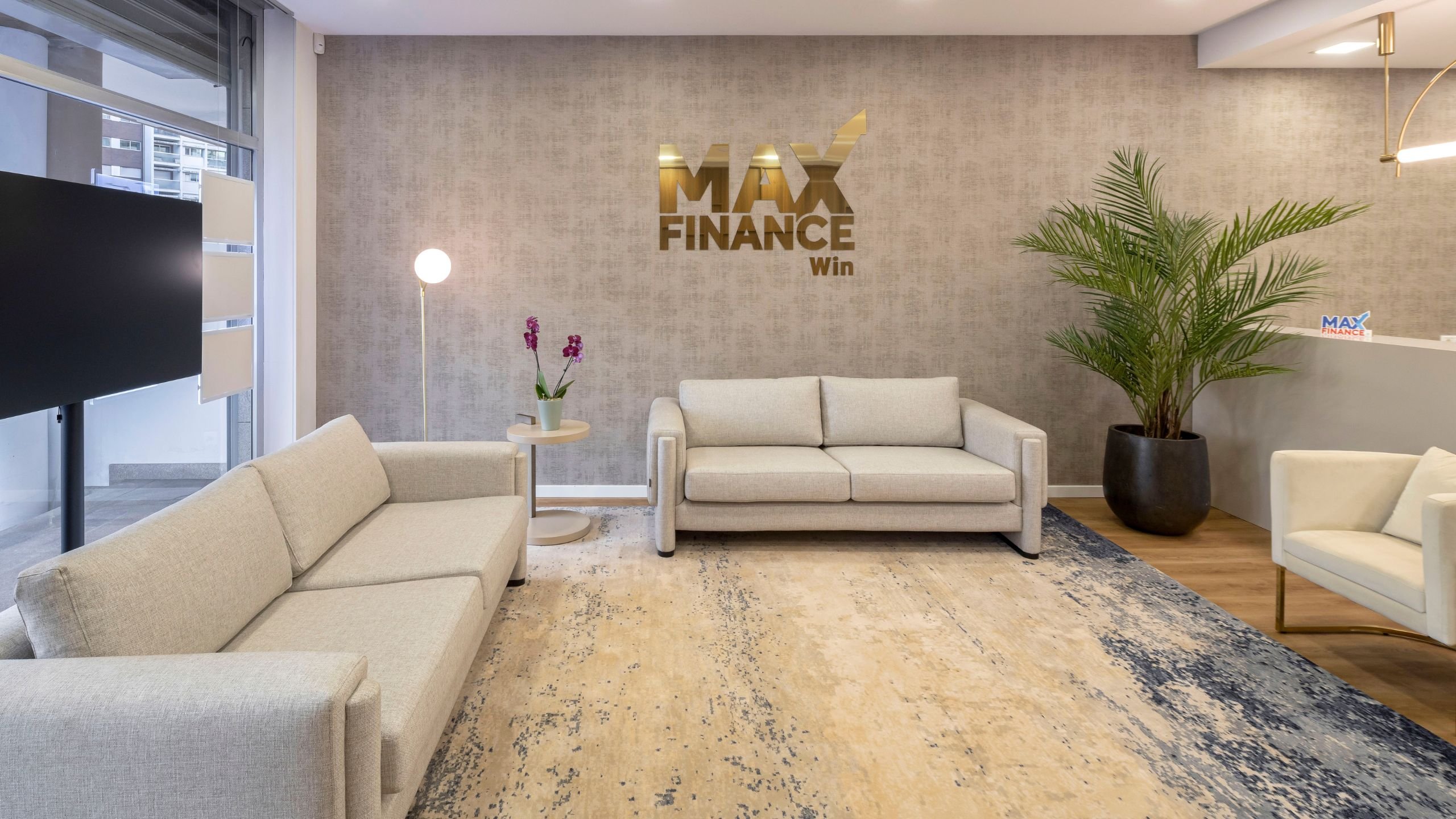 Perspetiva da sala de espera do escritório Max Finance Win, destacando os sofás em tom cru, a planta decorativa e o logotipo metálico dourado na parede principal.