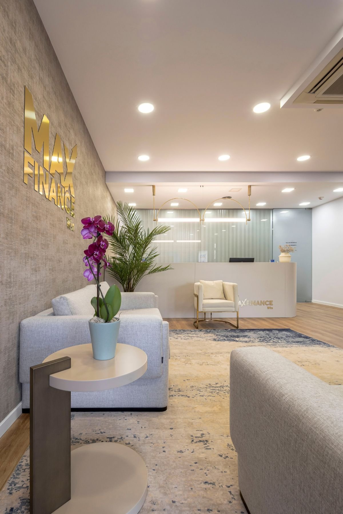 Vista lateral da entrada do escritório Max Finance Win, focando a mesa de apoio, a orquídea decorativa e a zona de espera luminosa.