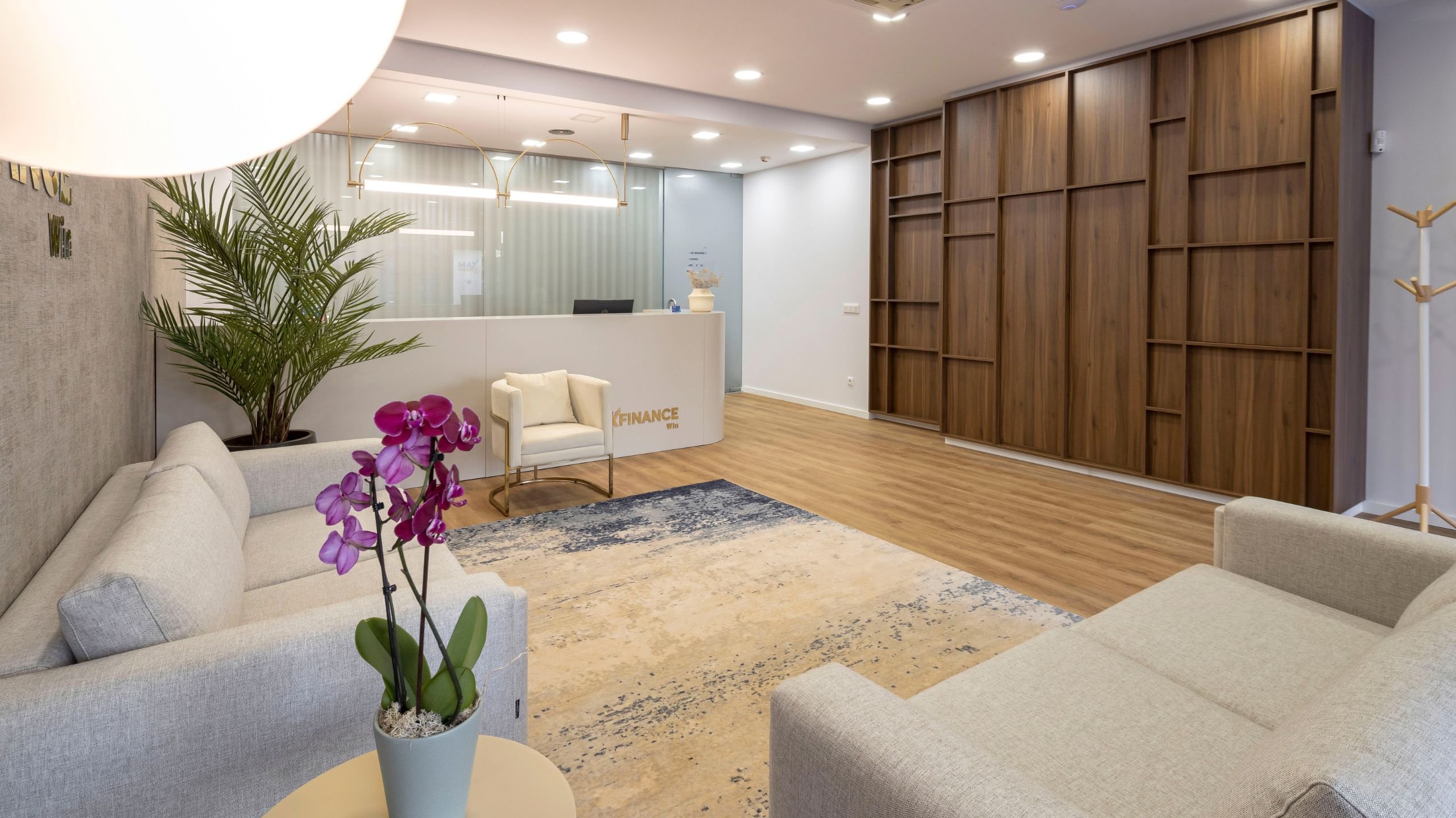 Visão ampla do hall de entrada da Imobiliária Max Finance Win, com destaque para a estante em madeira de nogueira mate, o balcão de receção, os sofás em tom cru e o tapete em tons claros e azul-marinho.