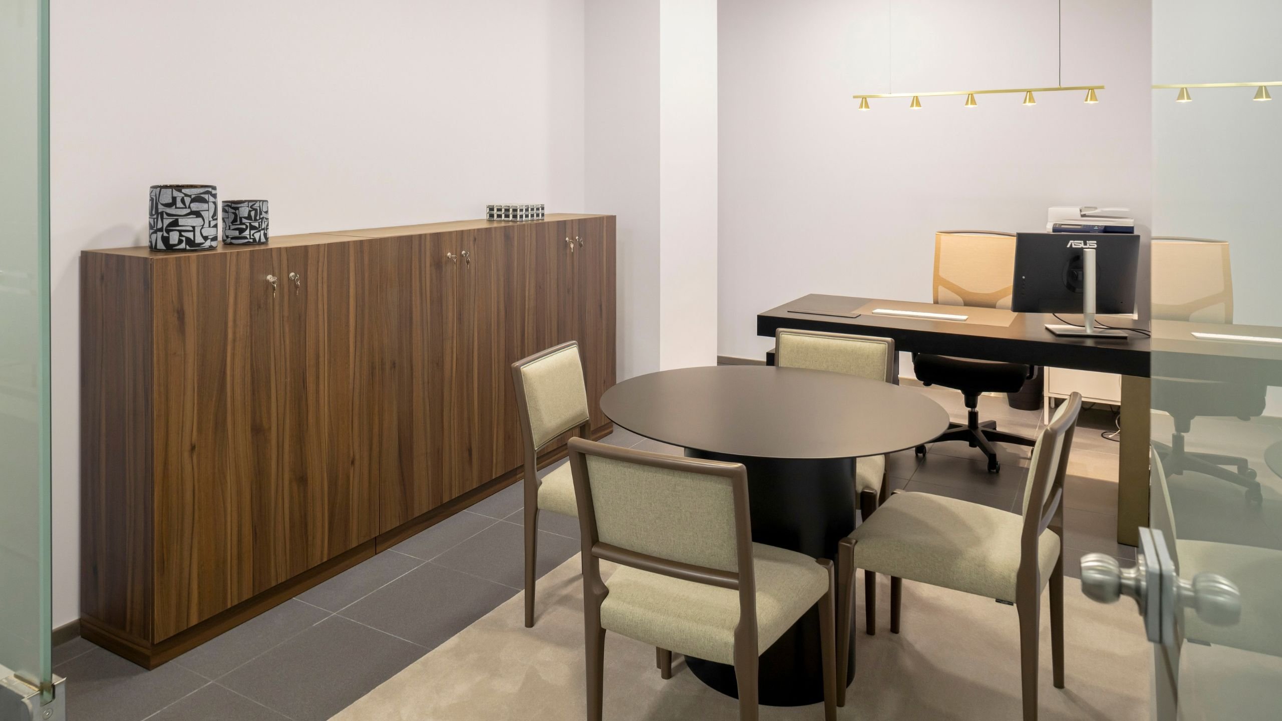 Visão geral do gabinete de direção da Max Finance Win, com mesa de reuniões redonda, cadeiras, secretária com módulo de arrumação e armários em nogueira mate.