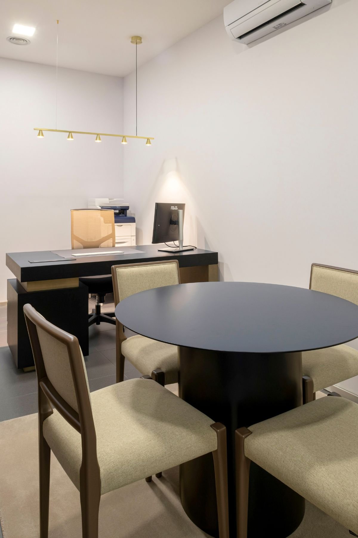 Perspetiva lateral da mesa de reuniões redonda em carvalho escurecido com cadeiras em primeiro plano e a secretária ao fundo.