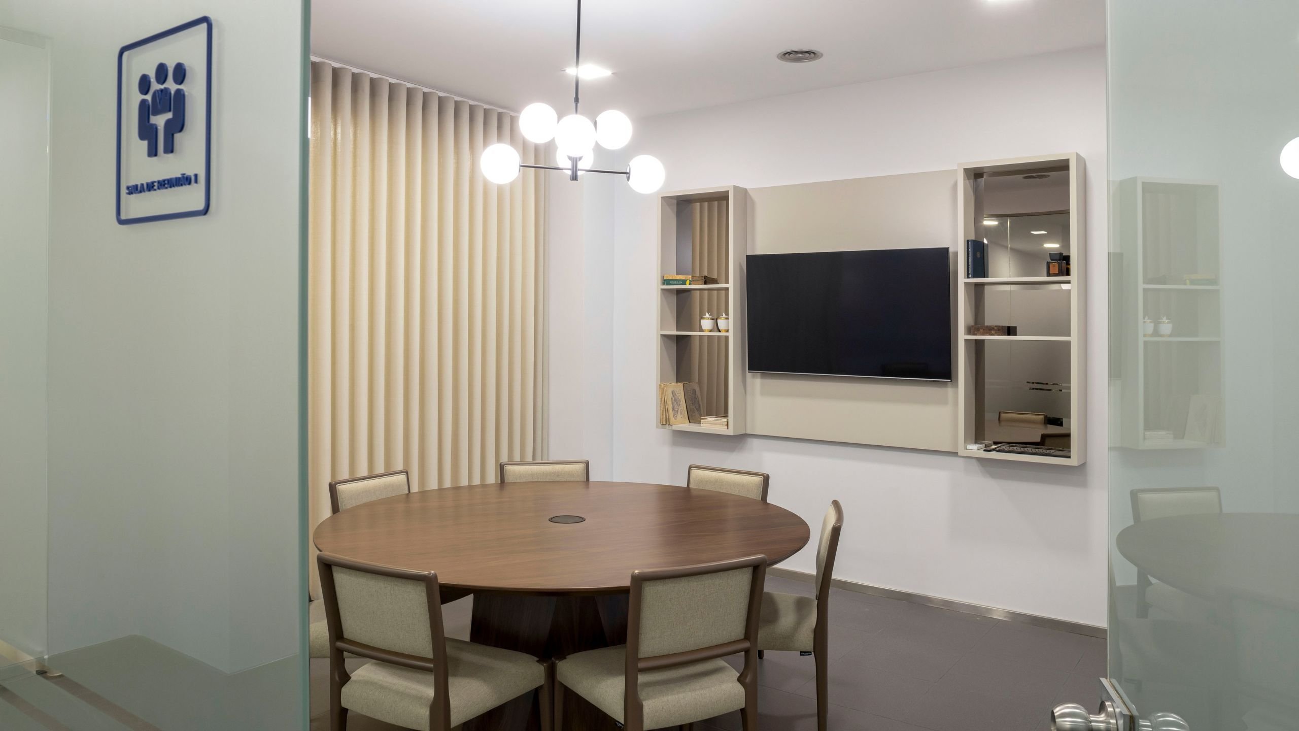 Visão geral da Sala de Reunião 1, com mesa redonda, cadeiras, estante suspensa em lacado bege para televisão e candeeiro de teto moderno de esferas.