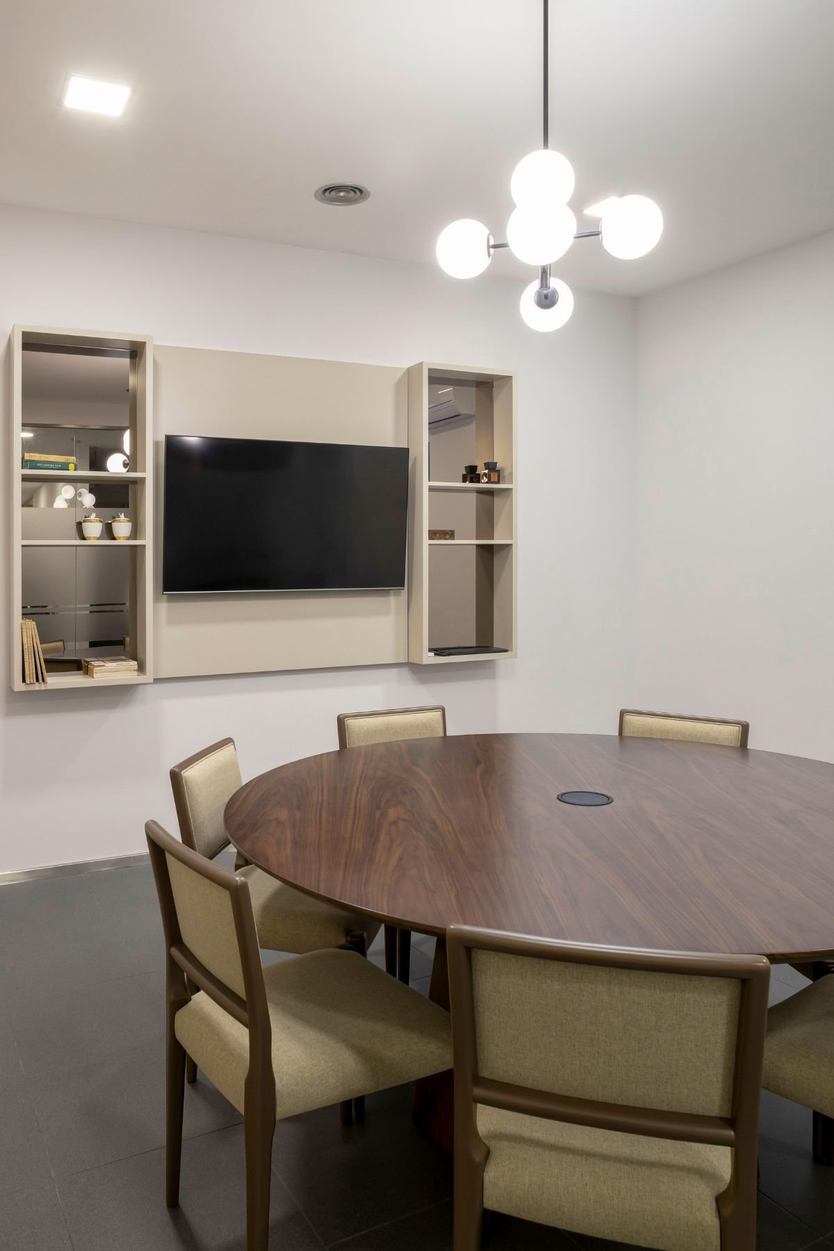 Vista angular da Sala de Reunião 1, focando o tampo em madeira da mesa, o conjunto de cadeiras e a estante de TV personalizada.