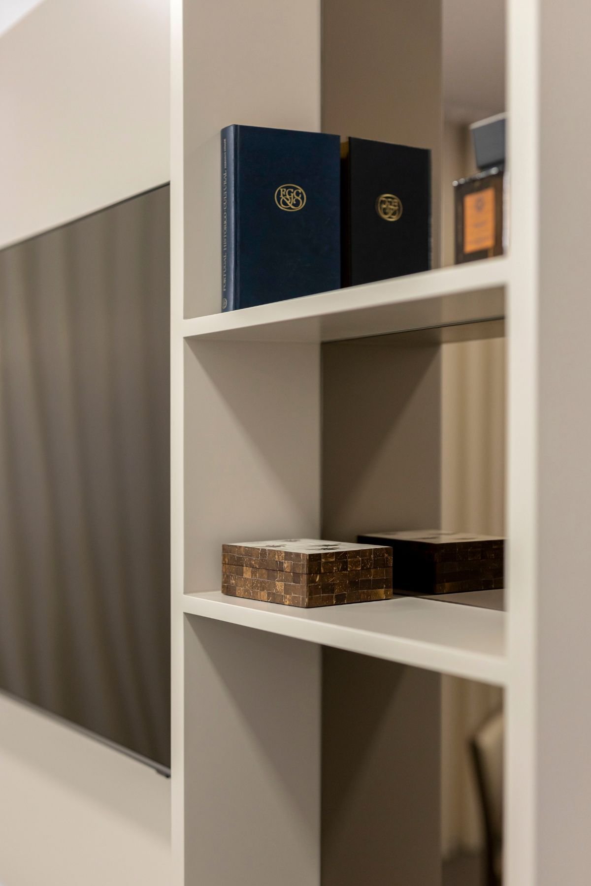 Pormenor da estante em lacado bege com fundo em espelho bronze, exibindo livros e peças decorativas organizadas nas prateleiras.