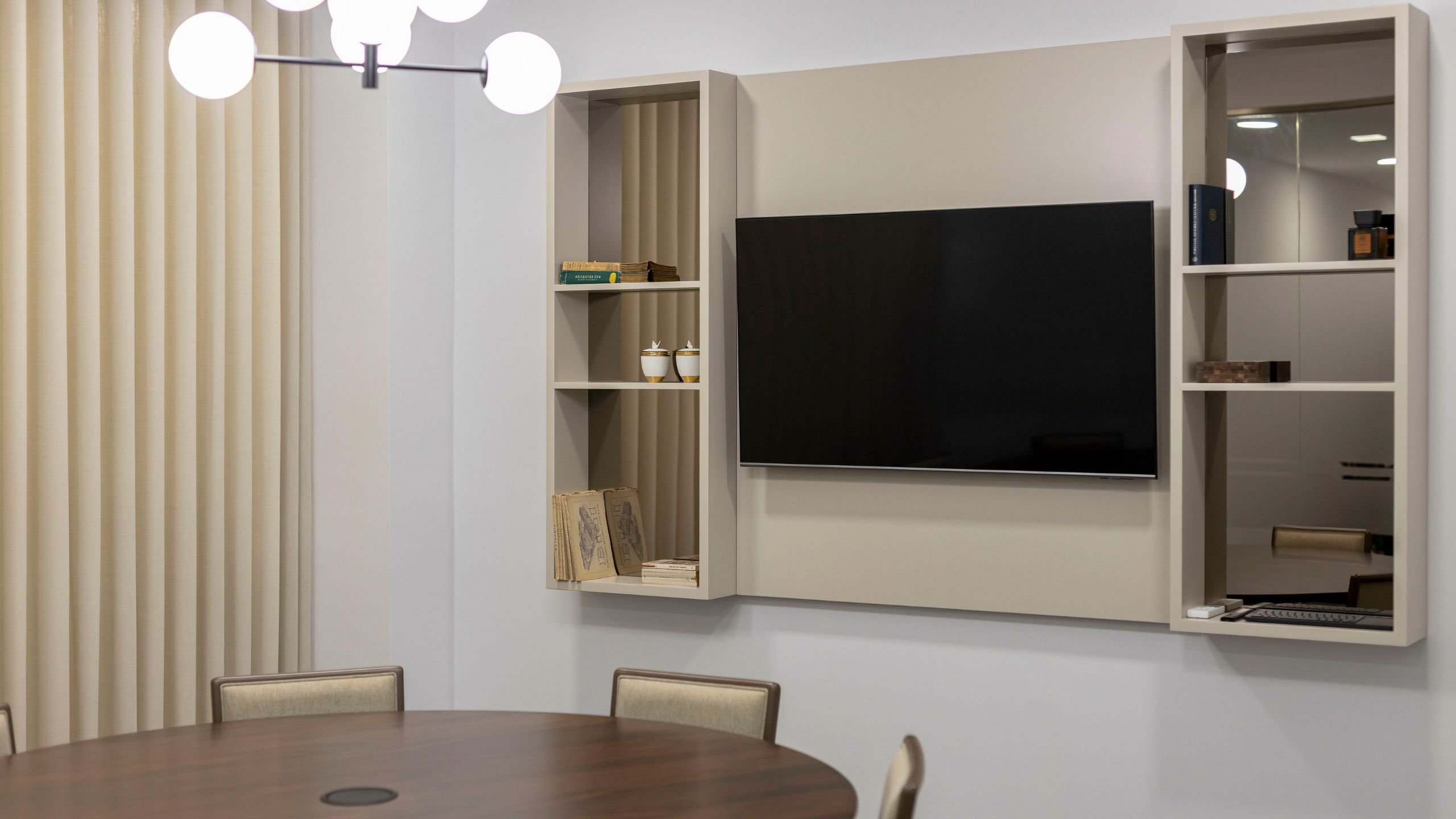 Plano aproximado da estante suspensa personalizada com televisão integrada, com a mesa de reunião redonda e cortinas neutras em segundo plano.
