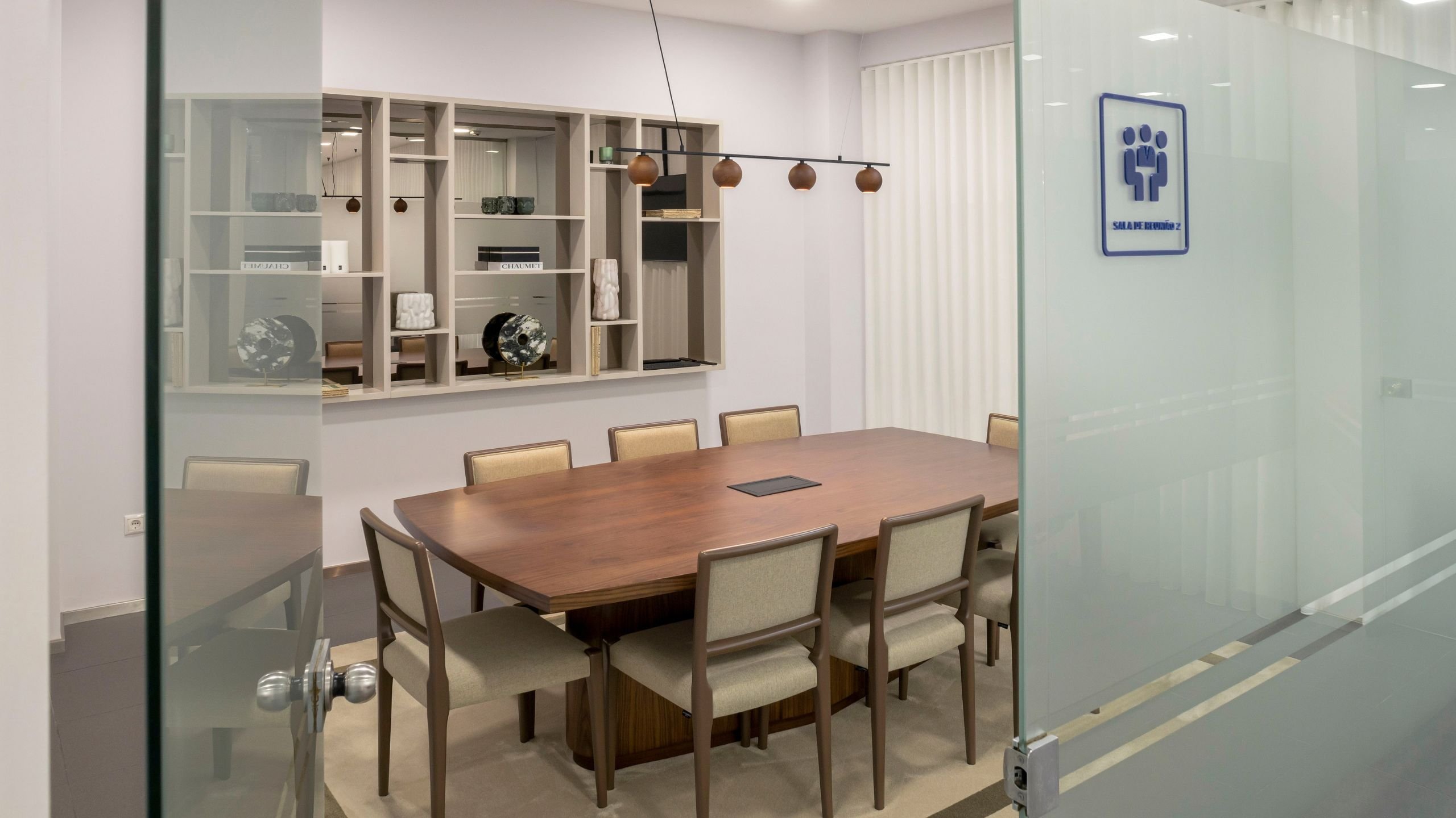 Visão geral da Sala de Reunião 2 através da porta de vidro, destacando a mesa retangular em nogueira mate, as cadeiras, a estante de parede suspensa e o candeeiro linear com focos em tons quentes.