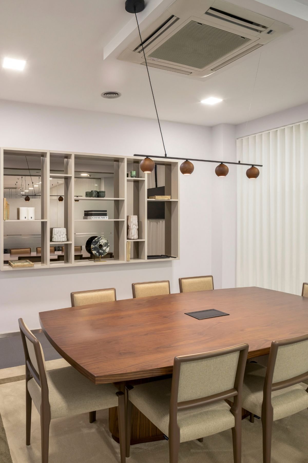 Perspetiva angular da mesa de reunião com o conjunto de cadeiras estofadas, enquadrando a estante em lacado bege e vidro bronze com peças decorativas ao fundo.