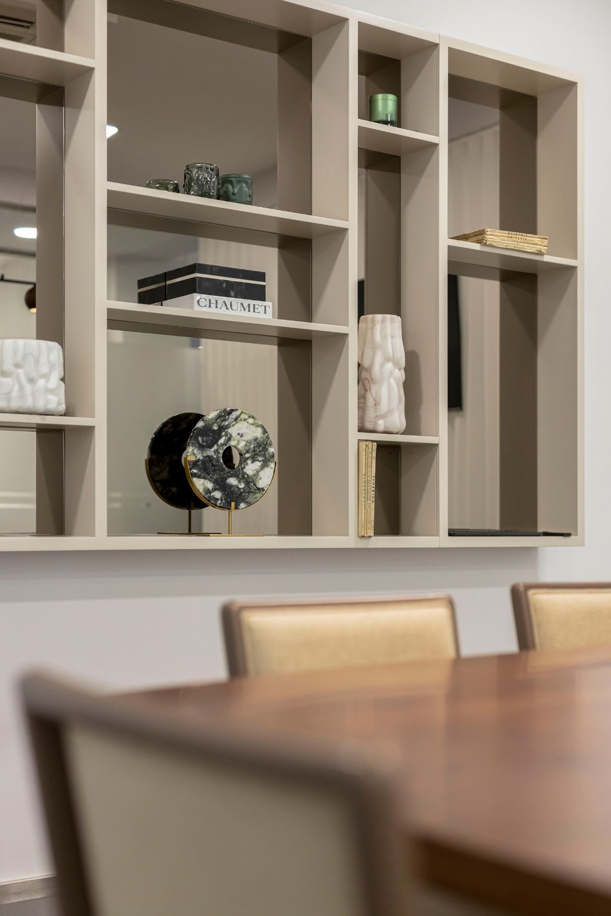 Detalhe da estante em lacado bege com acabamento em vidro bronze, exibindo livros de arte, peças em pedra decorativa e objetos de design sobre as cadeiras.