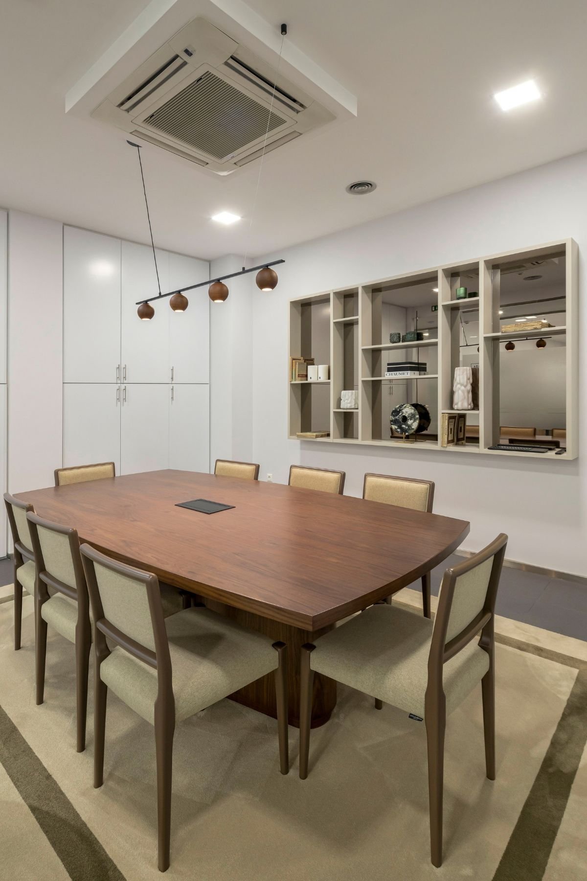 Vista ampla da Sala de Reunião 2, com foco na mesa de reuniões em nogueira para oito pessoas, o mobiliário embutido em lacado branco ao fundo e o candeeiro suspenso.