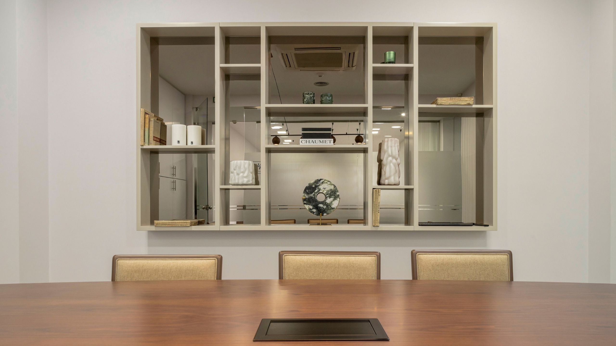Vista frontal enquadrada da estante suspensa em lacado bege e vidro bronze com nichos decorativos, posicionada acima da mesa de reunião em nogueira mate.