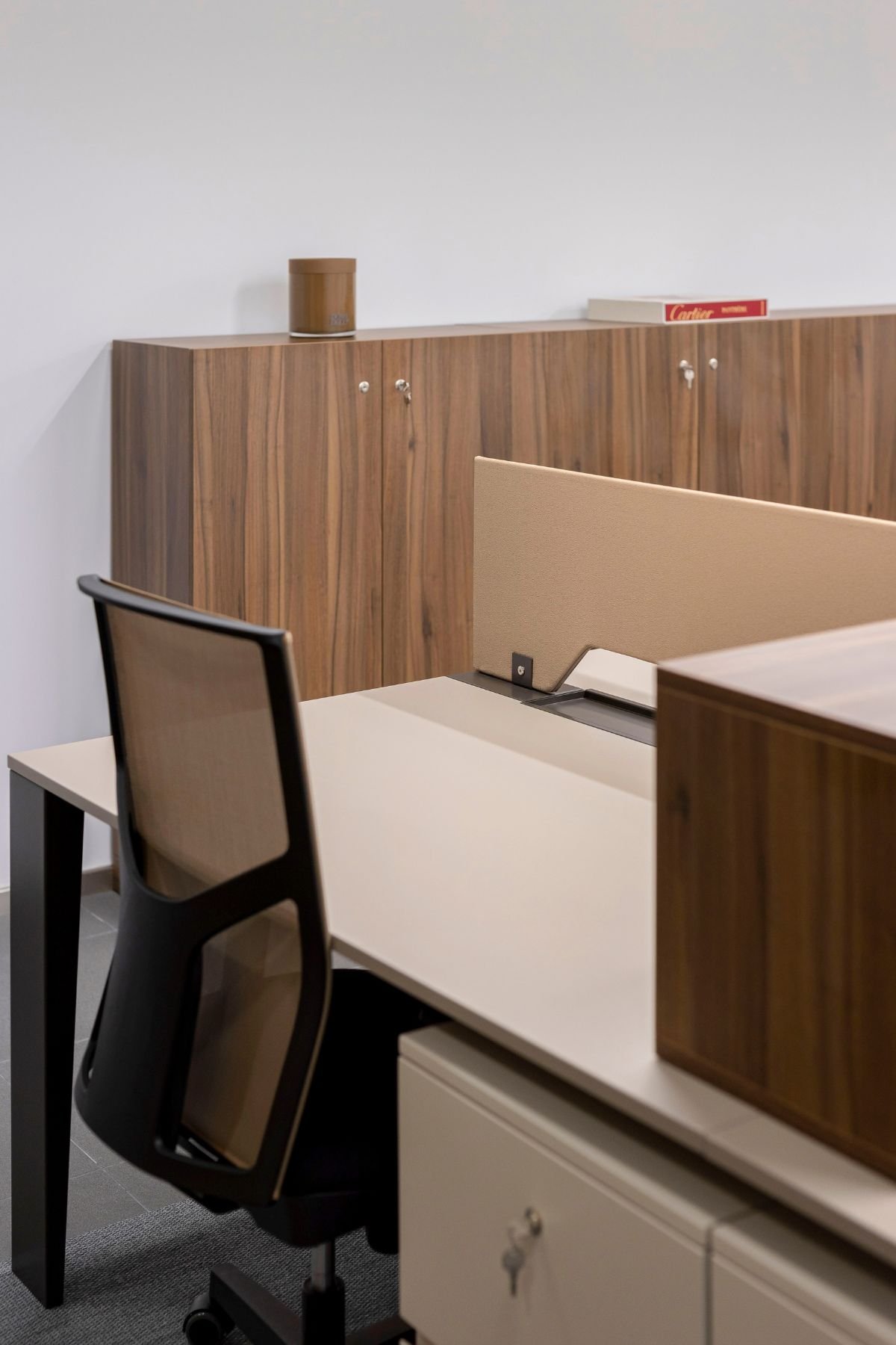 Detalhe em plano aproximado de uma workstation, com cadeira de escritório em rede, bloco de gavetas inferior com chave e armários corporativos em madeira ao fundo.