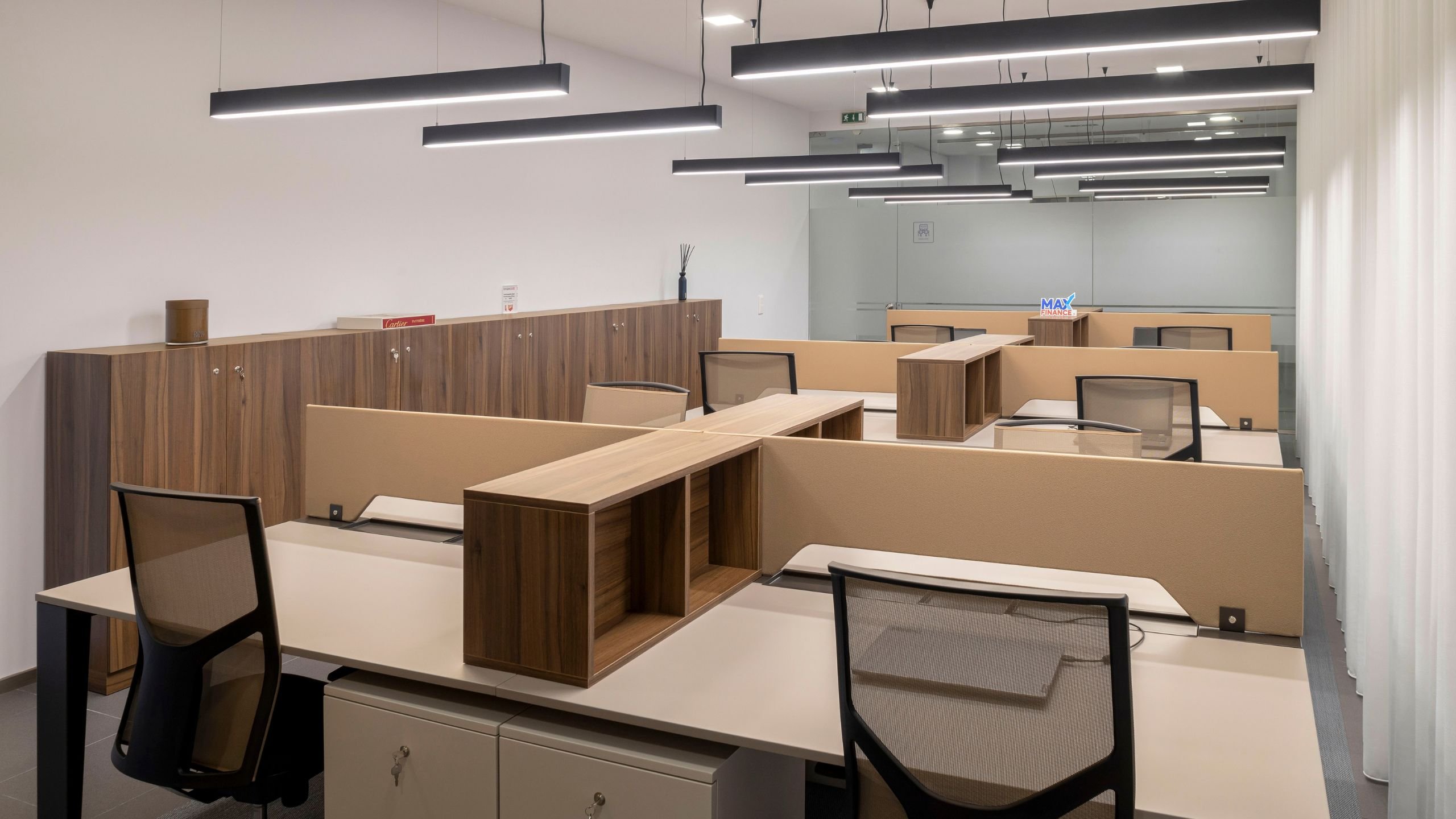 Perspetiva ampla do espaço de trabalho, mostrando a disposição modular das secretárias, os armários altos em tom de madeira ao longo da parede e a luminária suspensa sobre as bancadas.