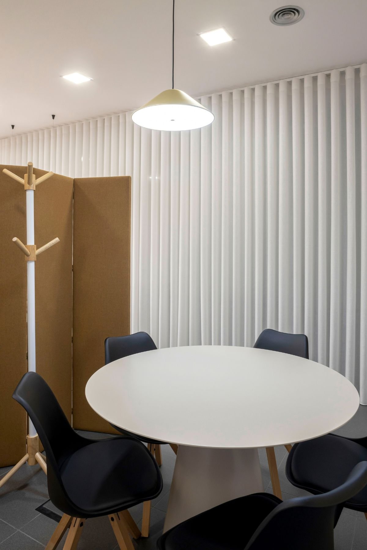 Área de reunião informal integrada no open space, composta por uma mesa redonda branca, cadeiras pretas com pés de madeira, um cabide de pé e um painel biombo divisório ao fundo.