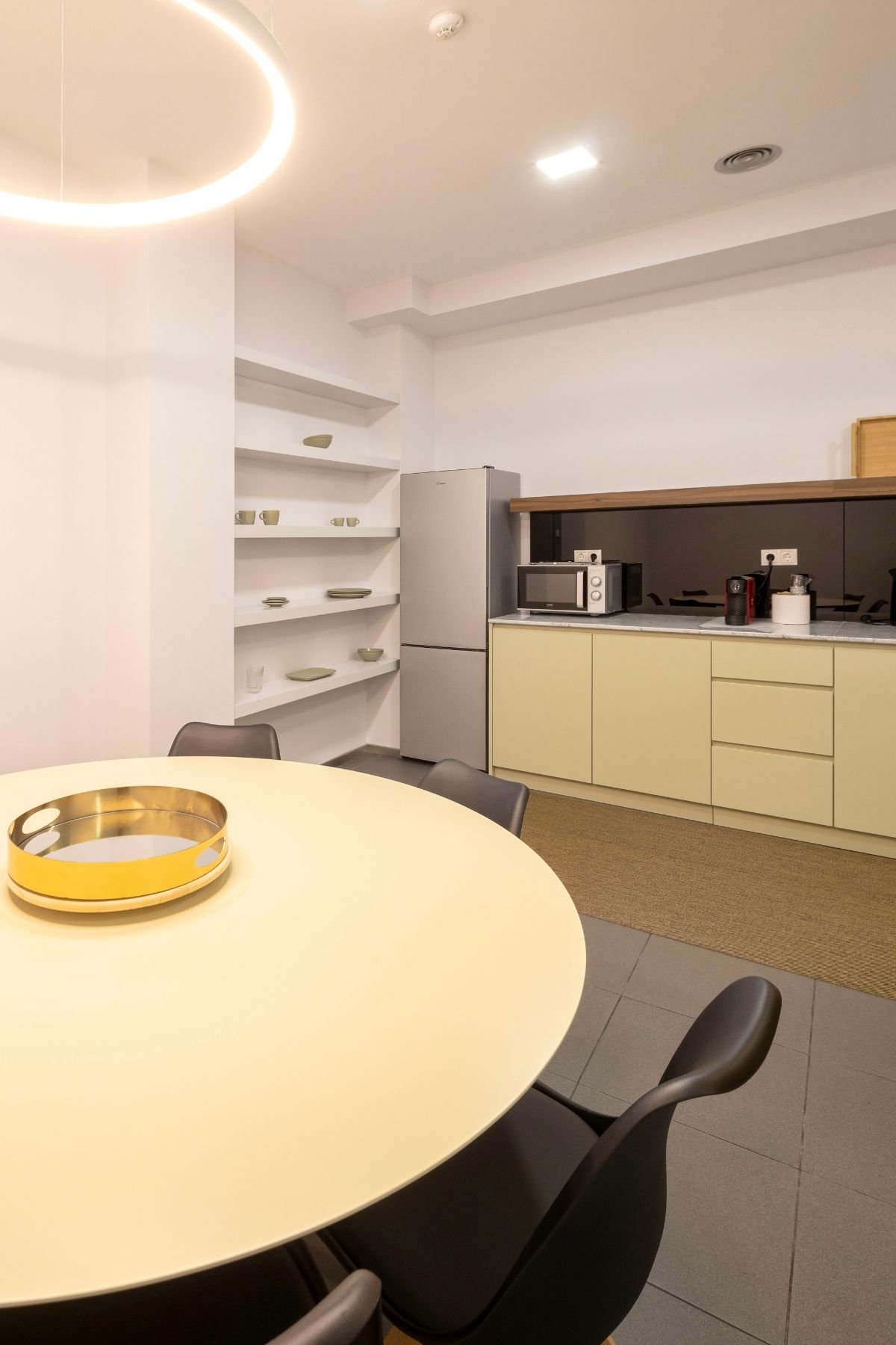 Perspetiva em primeiro plano da mesa redonda no espaço da copa, focando o tabuleiro decorativo dourado e as cadeiras pretas, com a zona de refeições e preparação ao fundo.
