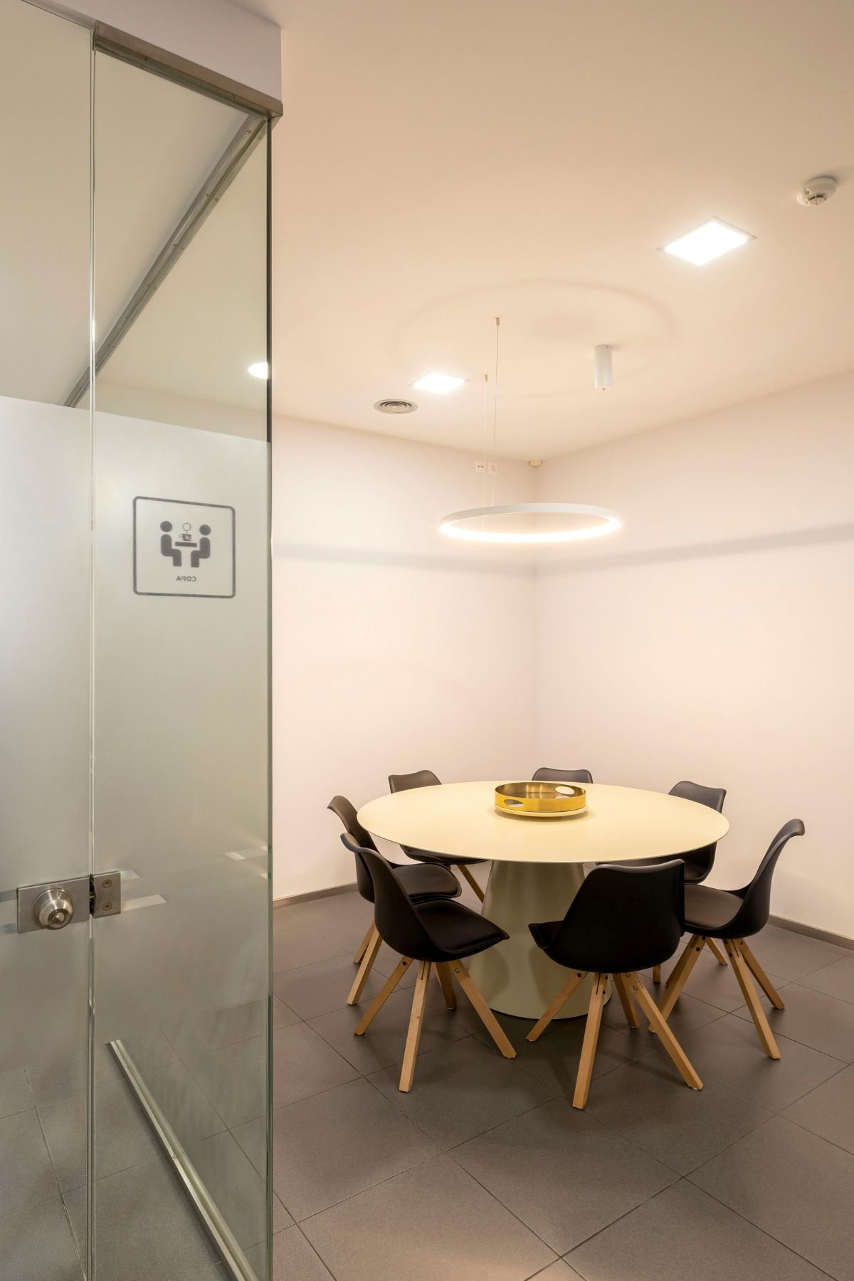 Vista em perspetiva da zona de refeições através da divisória de vidro da copa, apresentando a mesa cónica acompanhada por cadeiras pretas com pés em madeira e um candeeiro suspenso circular em anel.