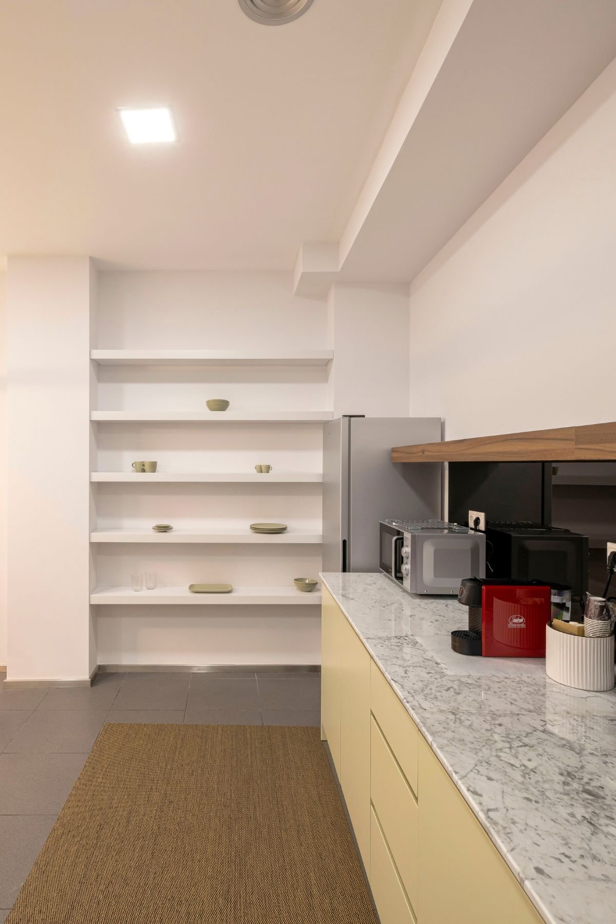 Pormenor do balcão da copa em mármore claro, evidenciando os eletrodomésticos de apoio, o revestimento escuro do backsplash e o alinhamento das prateleiras brancas ao fundo.