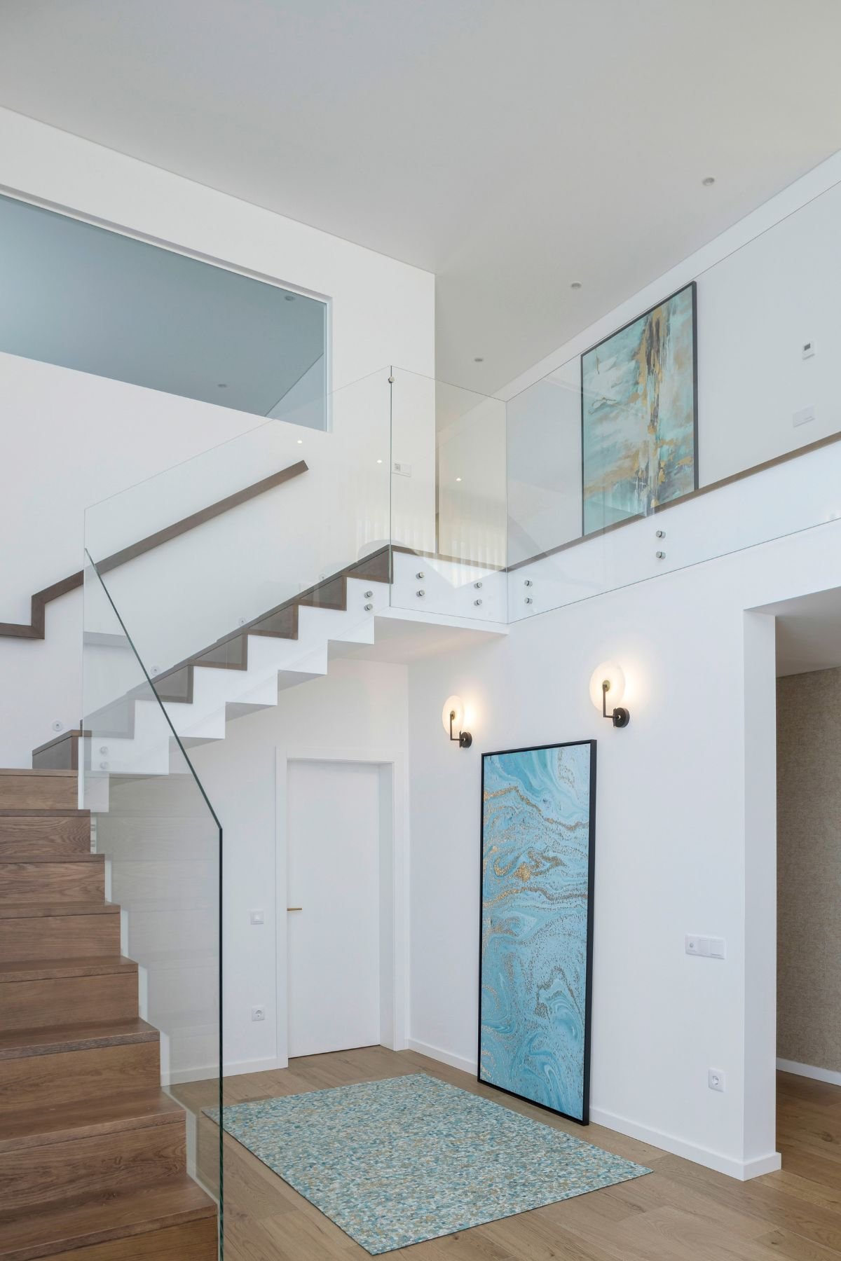 Hall de entrada amplo com escada em madeira e vidro, acompanhado por um quadro abstrato azul e apliques de parede num design de interiores elegante.