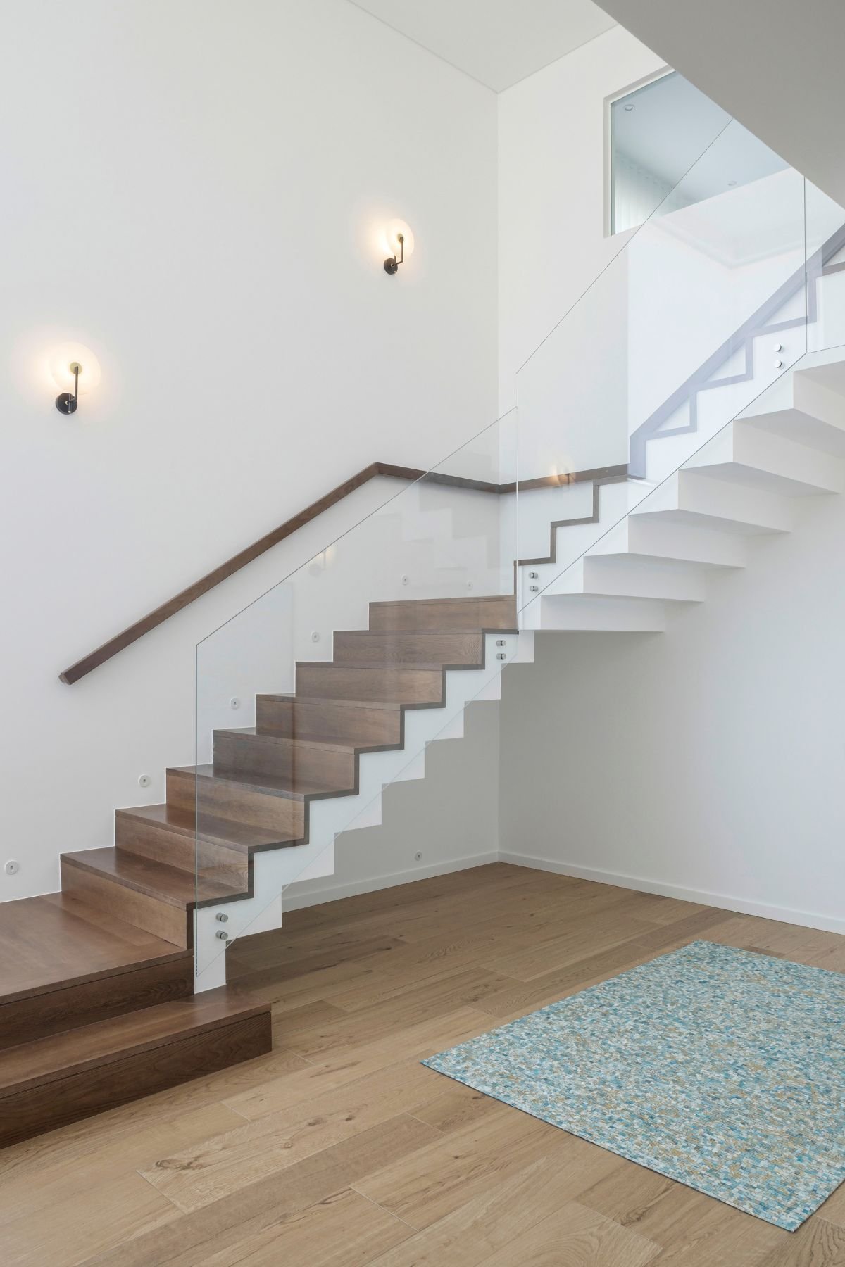 Pormenor da escada flutuante em madeira e estrutura branca com proteção em vidro e tapete decorativo no piso inferior.