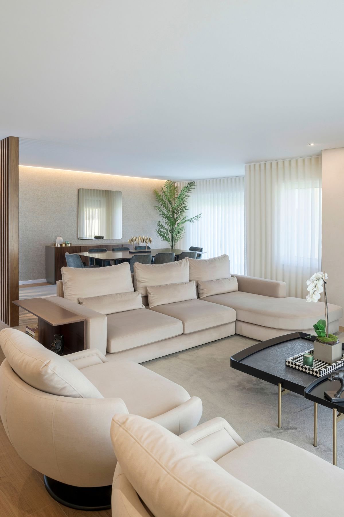 Perspetiva da sala de estar em plano aberto destacando o sofá com chaise longue e a ligação à sala de jantar ao fundo.