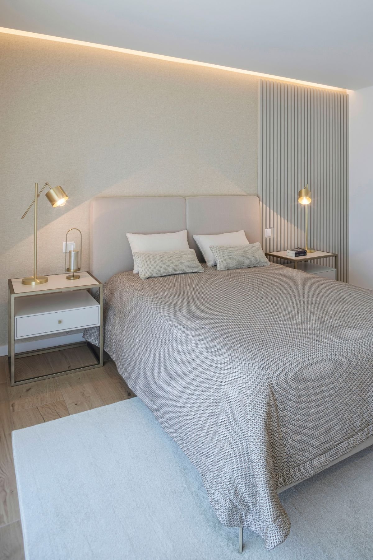 Perspetiva angular da suíte mostrando o painel ripado ao fundo e a cama de casal com roupa de cama bege.