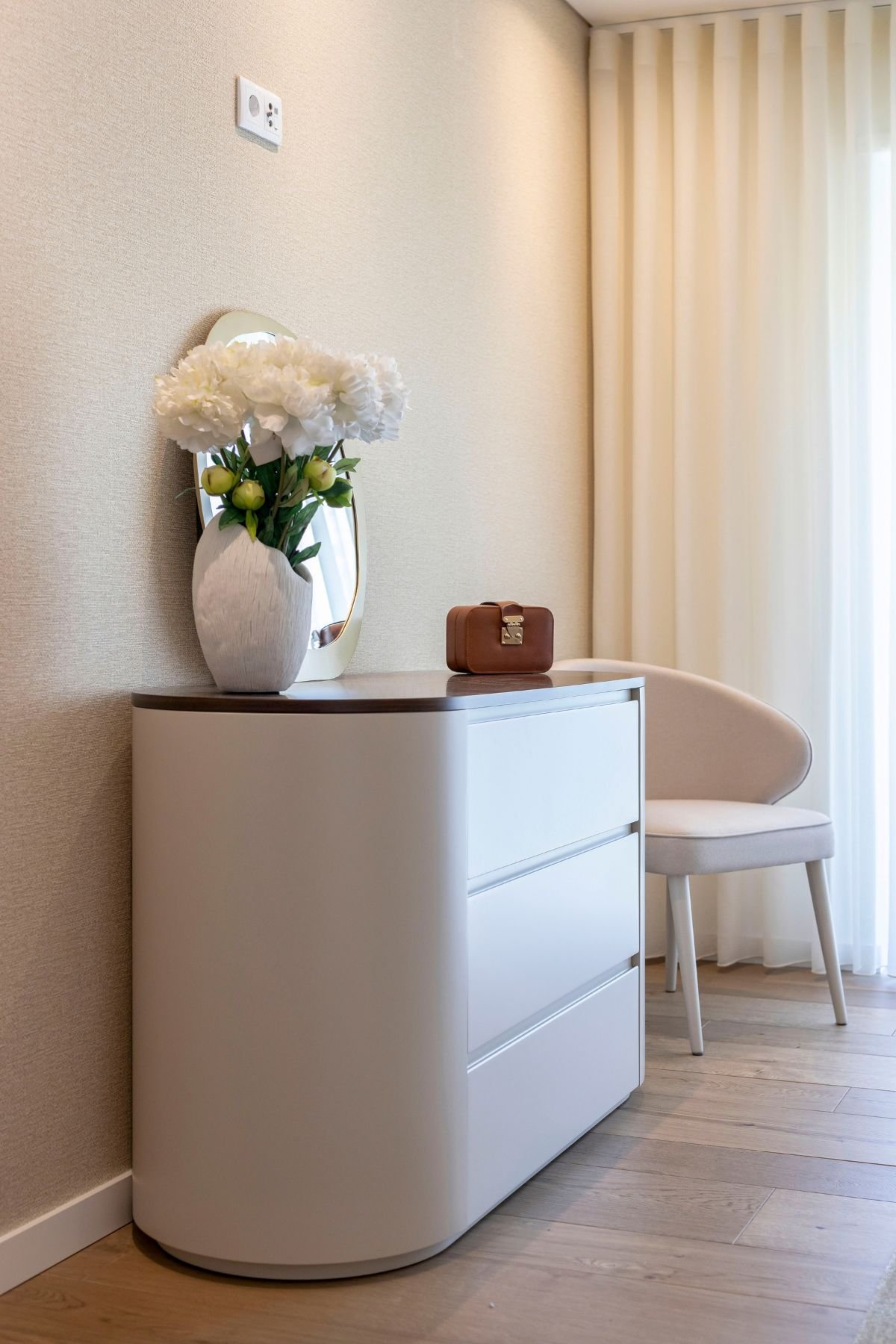 Ângulo aproximado da cómoda lacada com vaso de flores brancas e a cadeira Fortune ao fundo.
