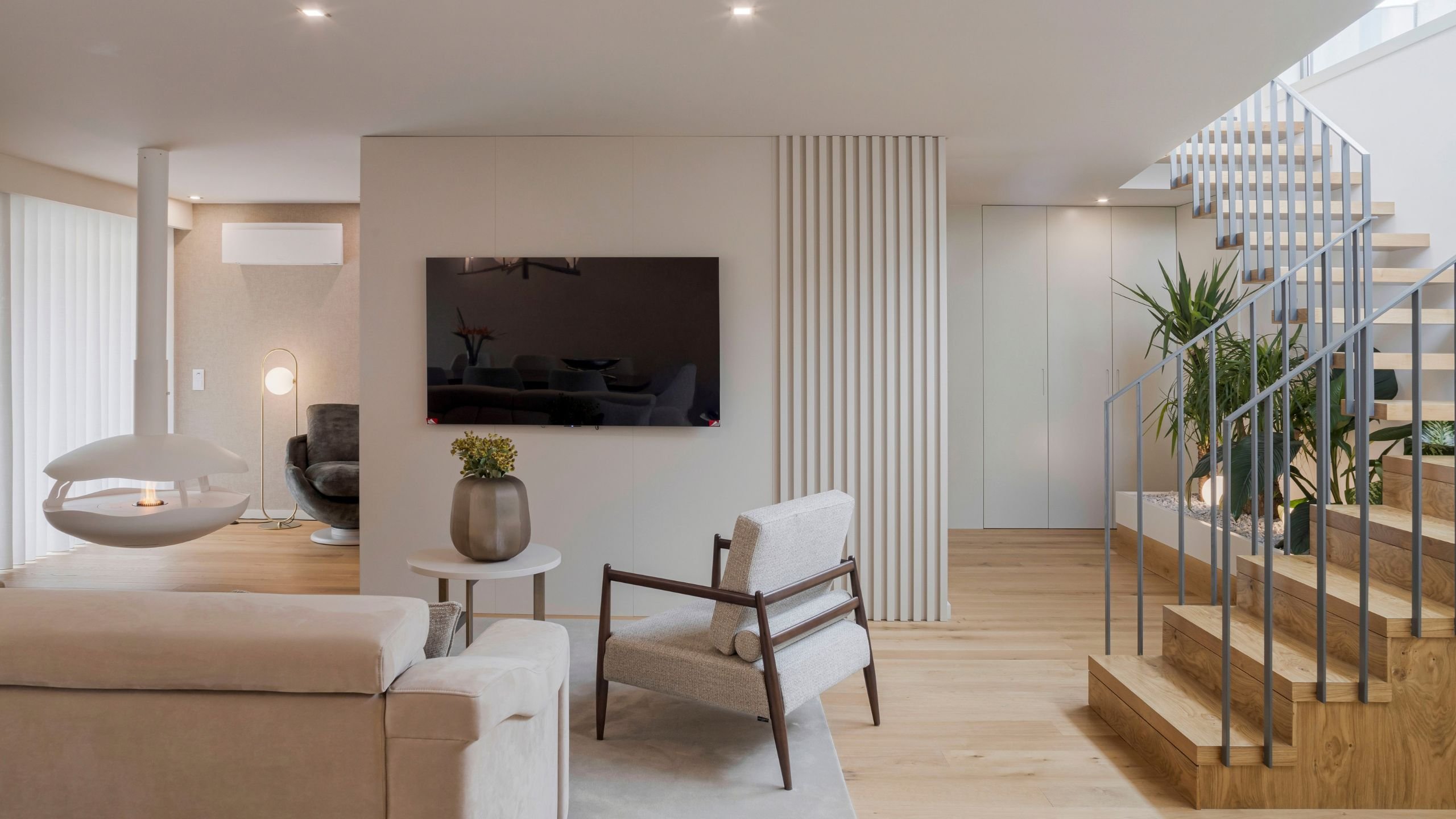 Zona de estar com lareira suspensa, escada em madeira e parede de ripado em open space sofisticado, espelho de excecional interior design e decoração em casas de luxo.
