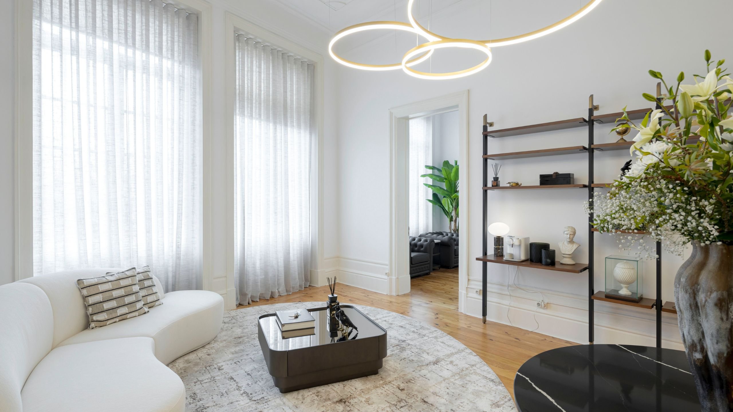 Sala de estar moderna de escritório com sofá curvo claro, mesa de centro espelhada, estante metálica e candeeiros de teto circulares num projeto de interiores harmonioso.