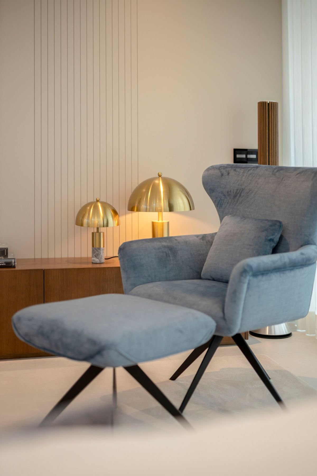 Cadeirão estofado em tecido azul com apoio de pés e candeeiro de mesa em dourado sobre o móvel tv na sala de estar moderna.